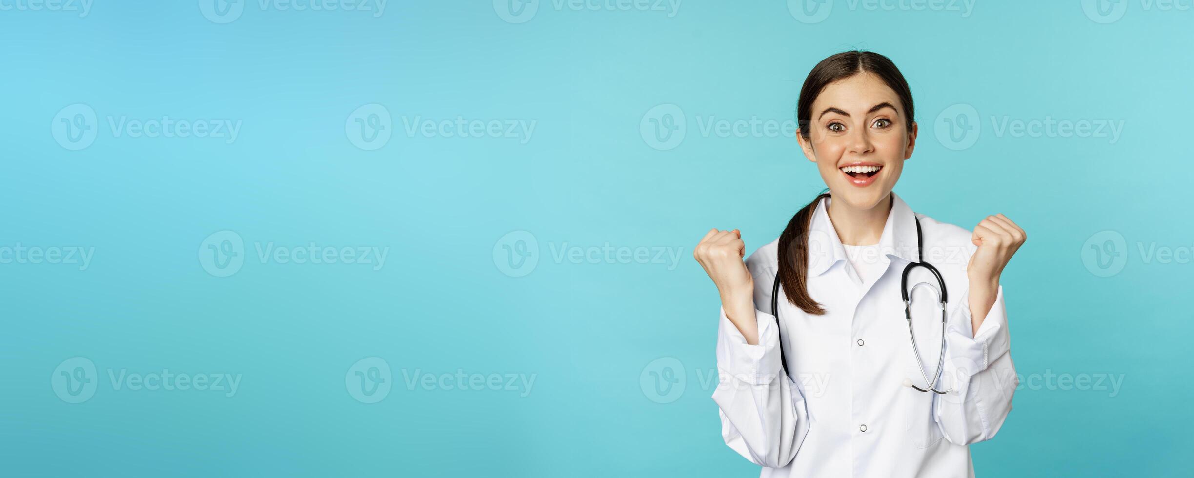Happy and excited young female doctor, physician celebrating, achieve goal and smiling pleased, winning, standing in white coat against blue background photo