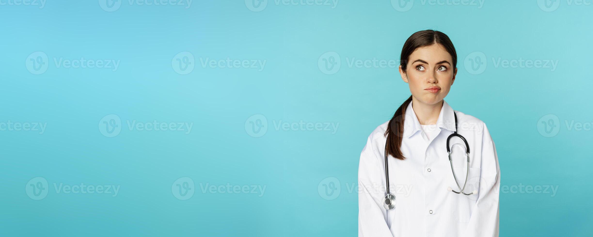 Image of woman doctor, female medial staff in white lab coat, looking away thoughful, making decision, thinking of smth, standing over blue background photo