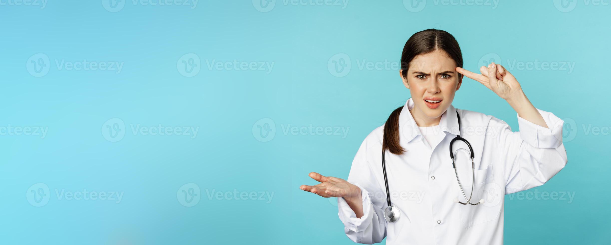 Annoyed woman doctor pointing finger at head, scolding someone stupid, crazy or strange, standing over torquoise background photo