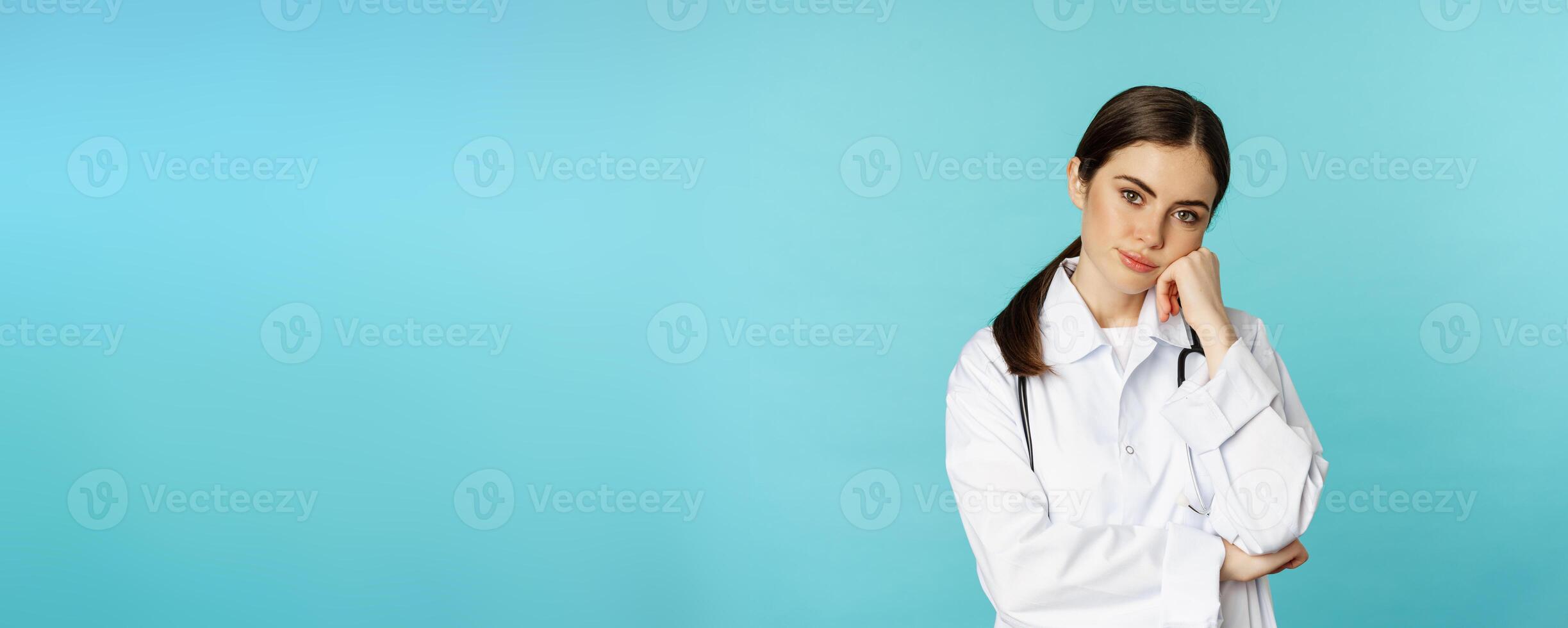 Thoughtful medical worker, young doctor woman looking unamused, bored, standing in white coat against blue background photo