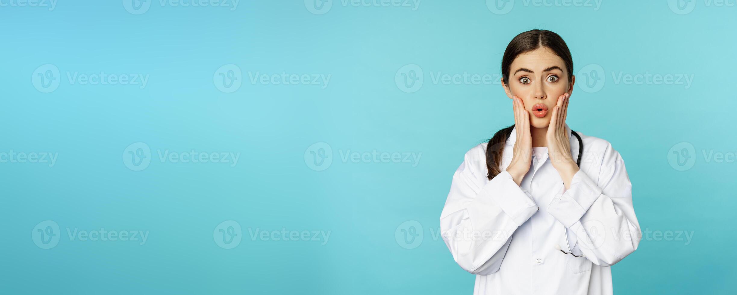 Portrait of shocked female doctor, worried healtcare worker in white robe, looking concerned at patient, standing over blue background photo
