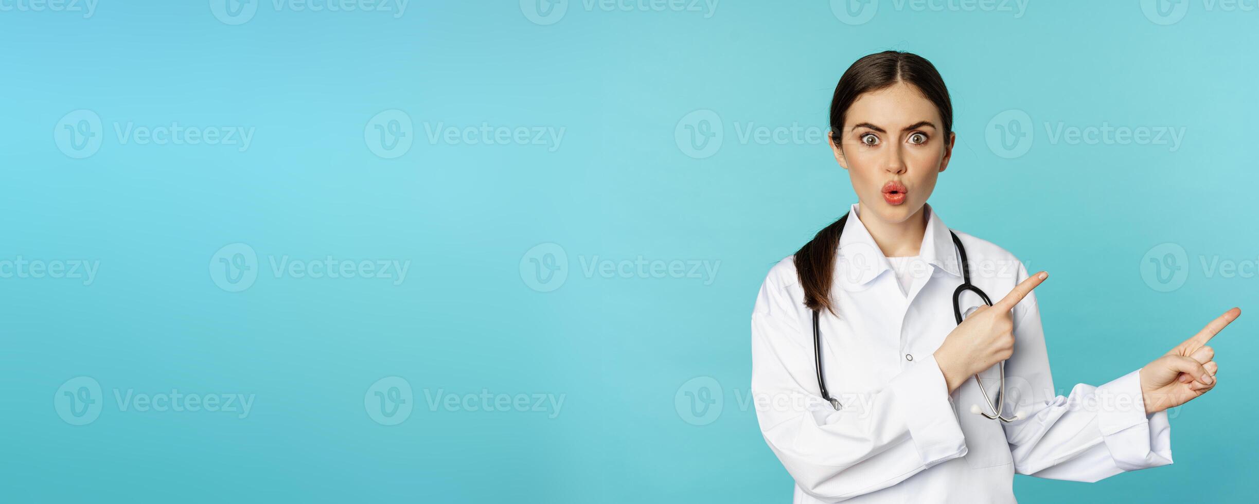 Portrait of surprised medical worker, woman doctor pointing fingers right and say wow amazed, reacting interested, standing over torquoise background photo