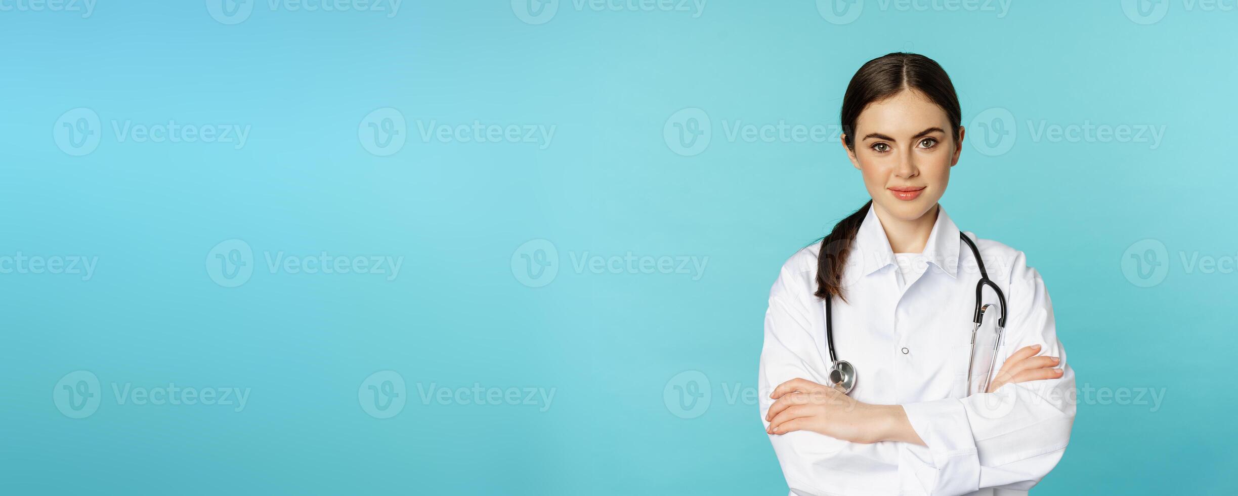 Medical staff and doctors concept. Young smiling female doctor, healthcare worker in white coat and stethoscope, looking confident, waiting patients, blue background photo