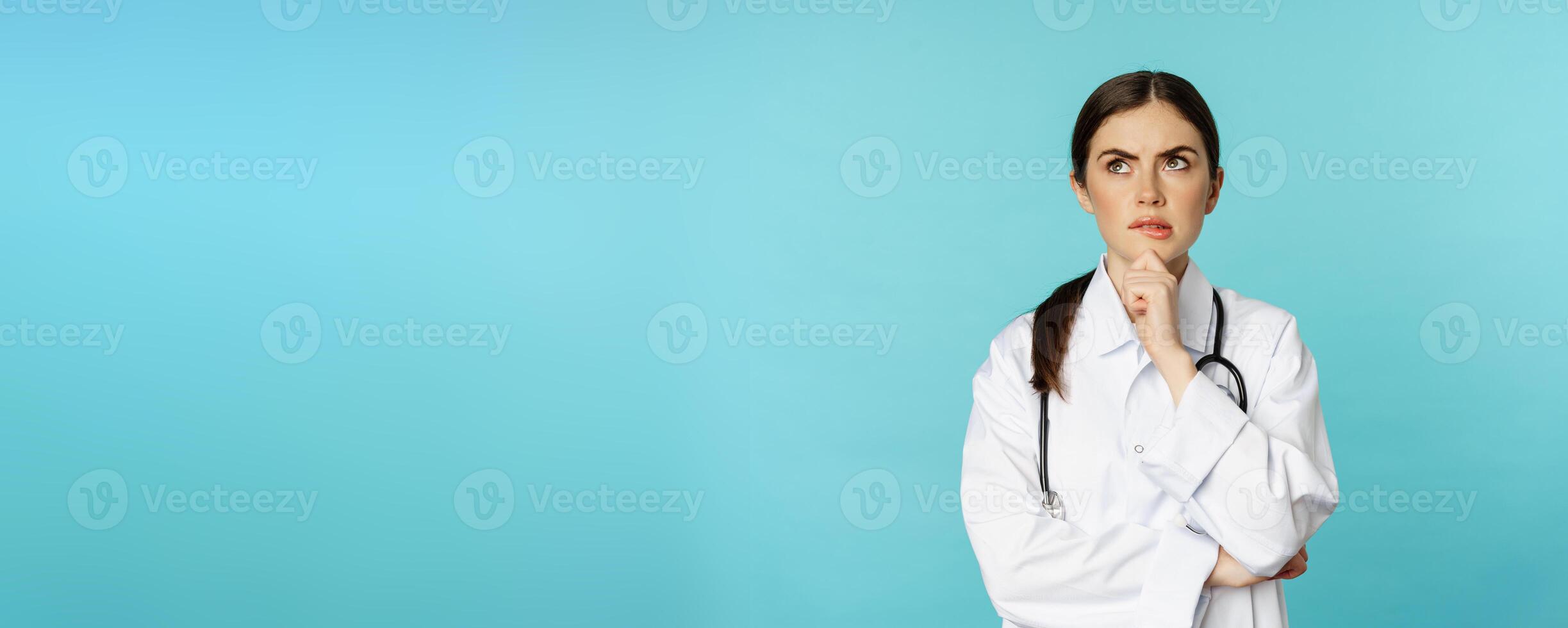 Young female doctor, hospital worker in white coat, thinking and looking away thoughtful, searching solution, standing over toquoise background photo