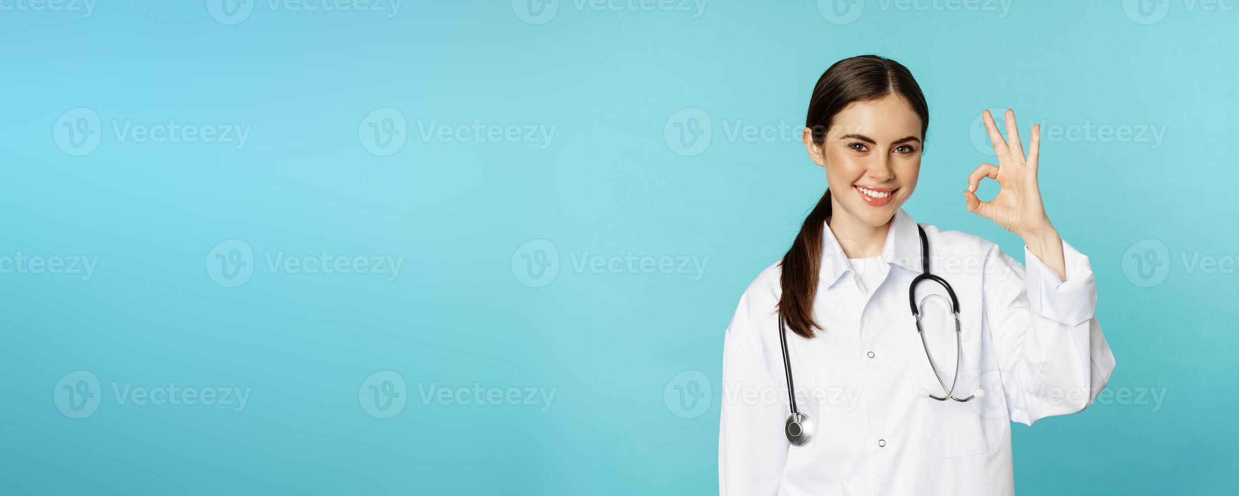 Portrait of satisfied, smiling medical worker, woman doctor showing okay, ok, zero no problem gesture, excellent sign, standing pleased over torquoise background photo