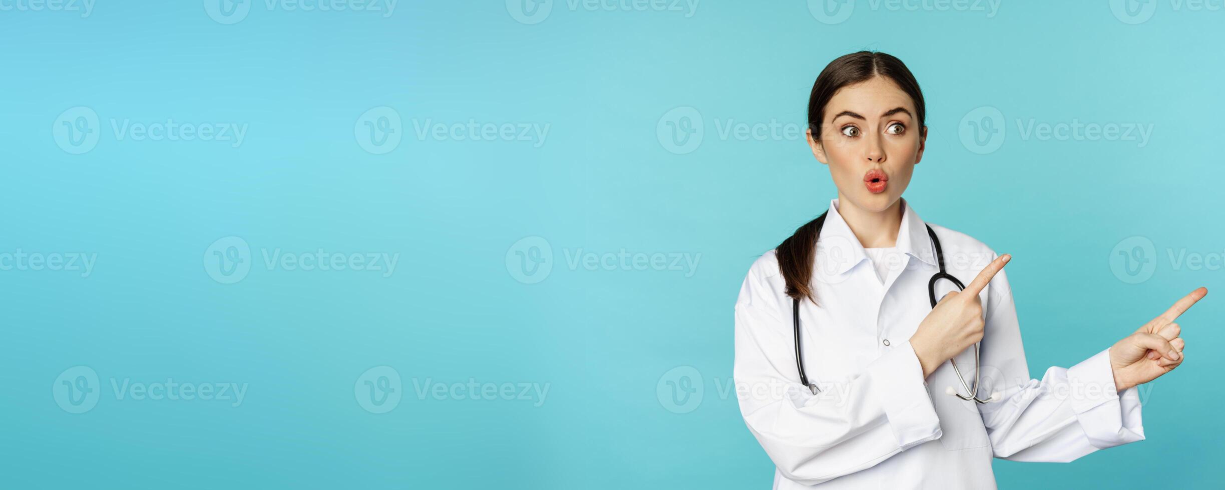 Portrait of surprised medical worker, woman doctor pointing fingers right and say wow amazed, reacting interested, standing over torquoise background photo