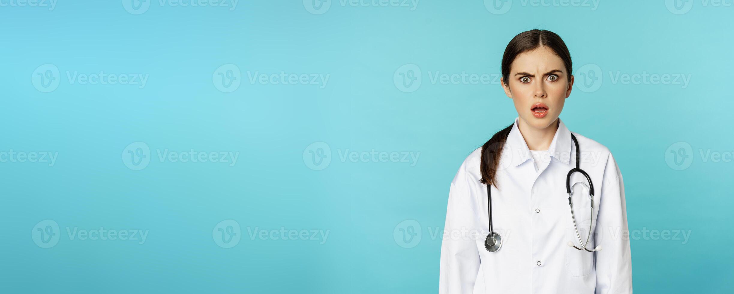 Portrait of shocked woman doctor, female hospital intern in white coat, looking concerned and confused at camera, disbelief face, standing over torquoise background photo
