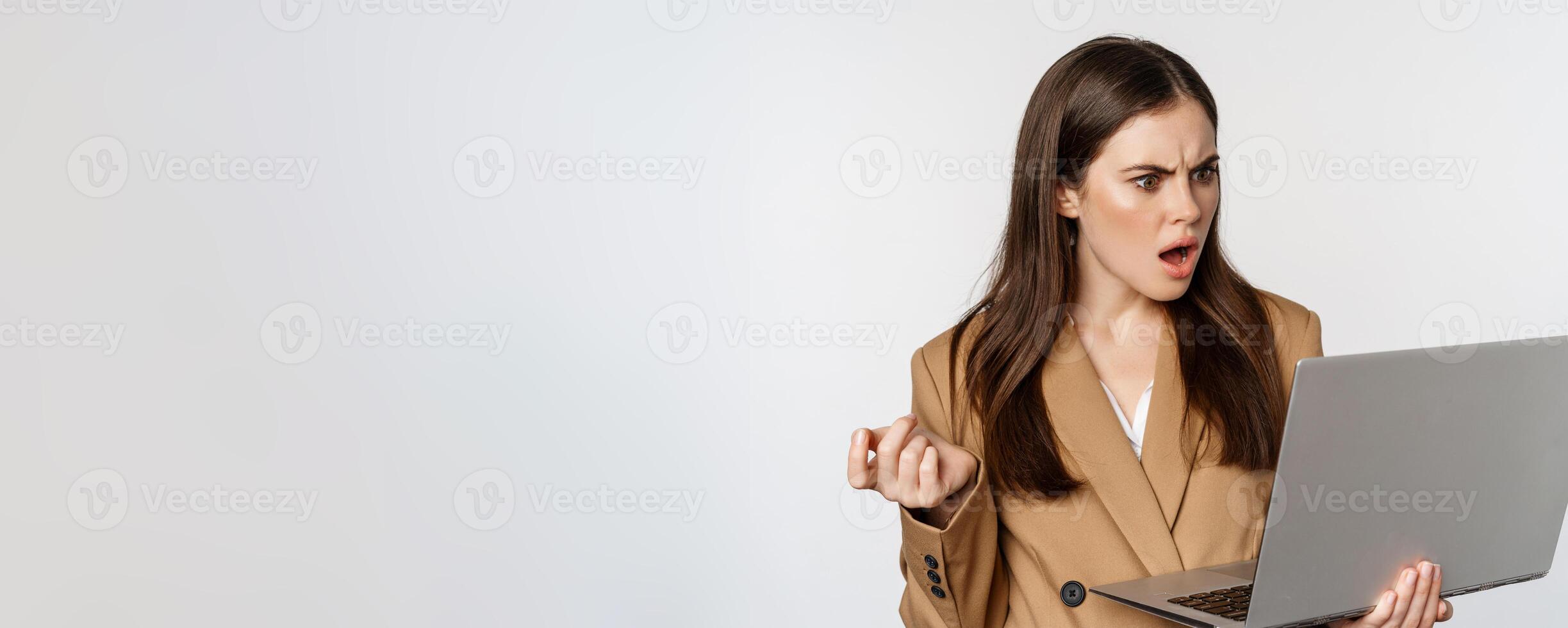 Shocked and disappointed business woman looking at laptop screen with offended, frustrated face expression, standing against white background photo