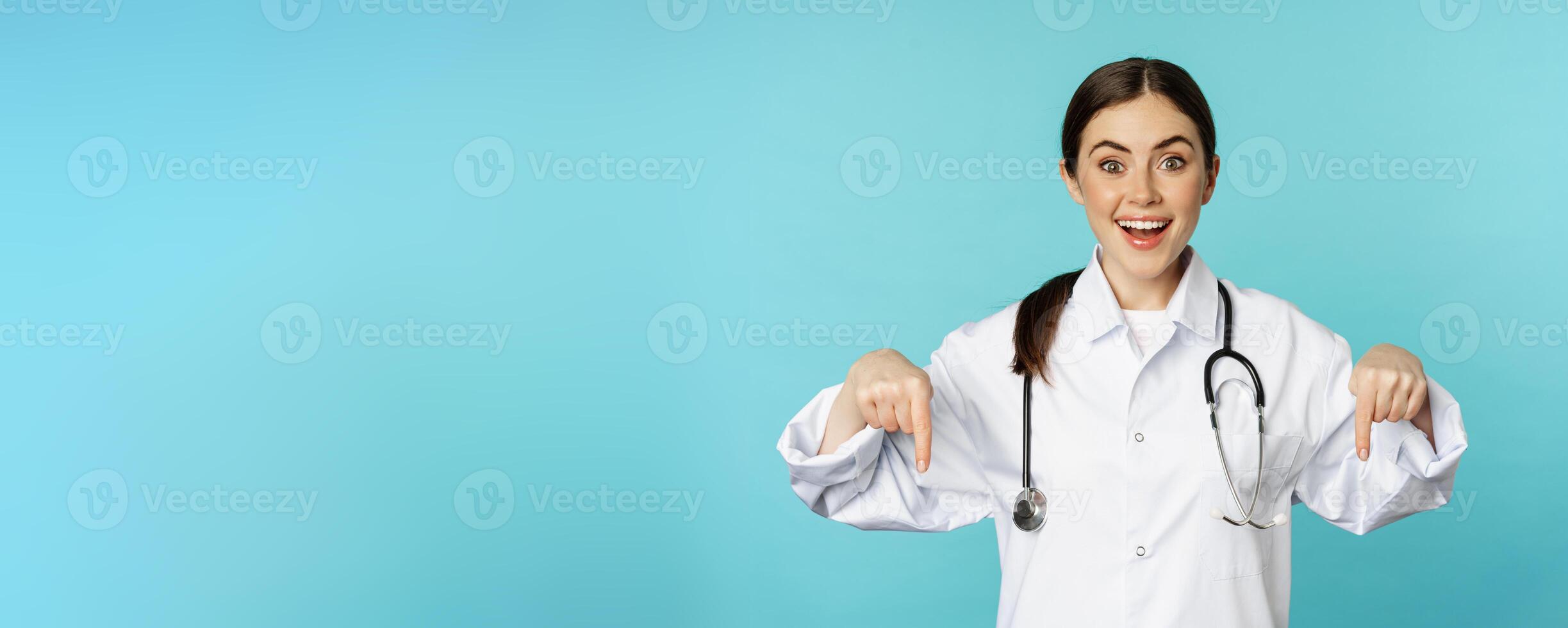 Enthusiastic healthcare worker, young woman doctor in white coat, pointing fingers down and smiling, showing pharmacy advertisement, medical promo, blue background photo