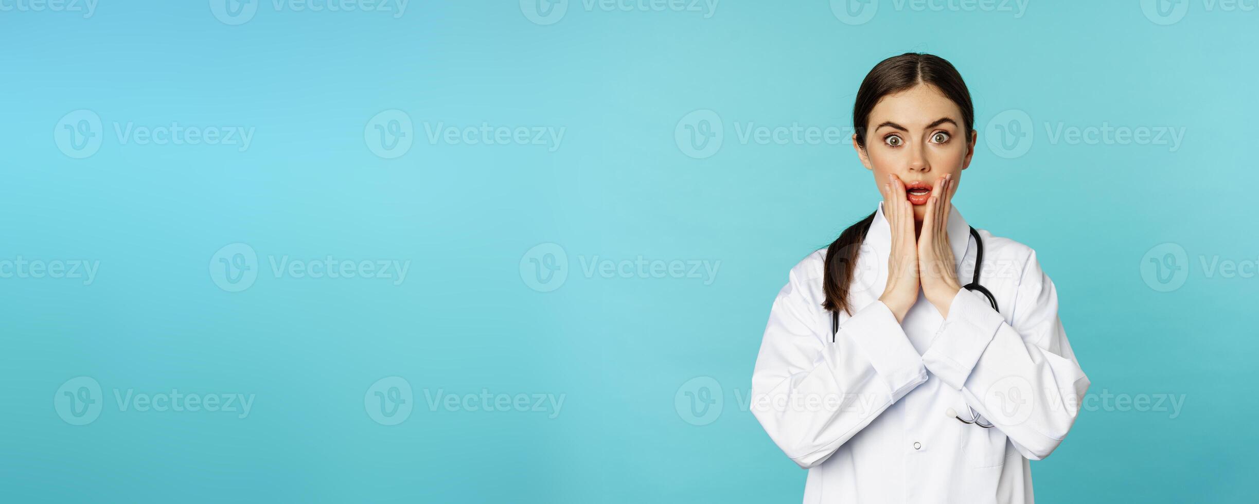 Portrait of shocked woman doctor, female hospital intern in white coat, looking concerned and confused at camera, disbelief face, standing over torquoise background photo