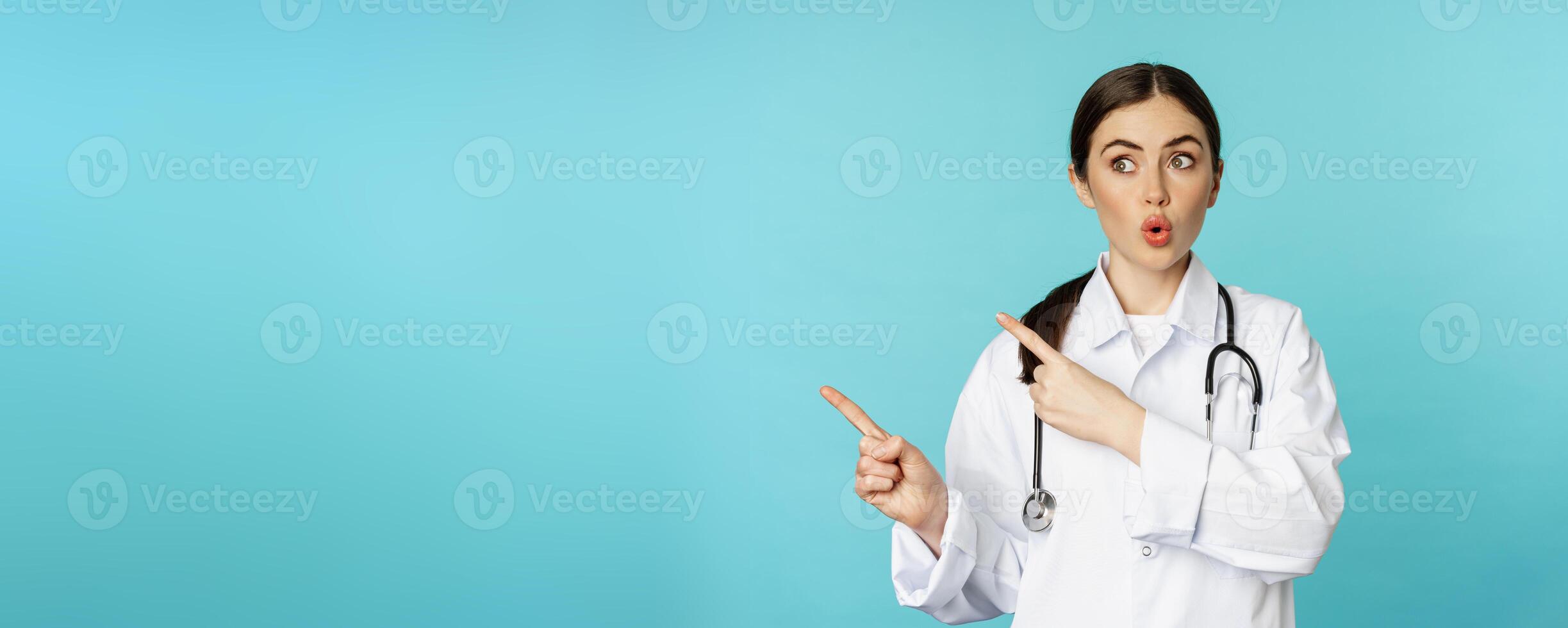 Surprised female doctor, physician with stethoscope, pointing and looking left with amazed, wow face, standing over torquoise background photo