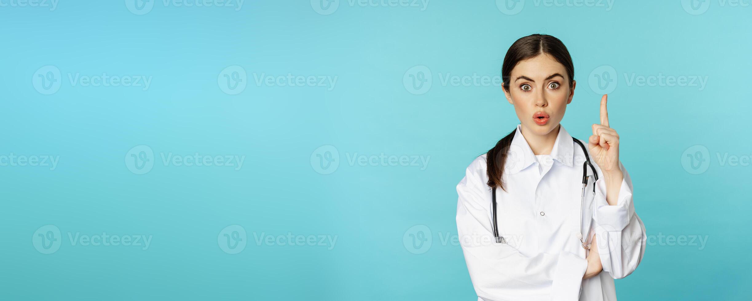 Young doctor, woman physician in white coat raising finger, pointing up, suggesting smth, has solution, revelation, standing over torquoise background photo
