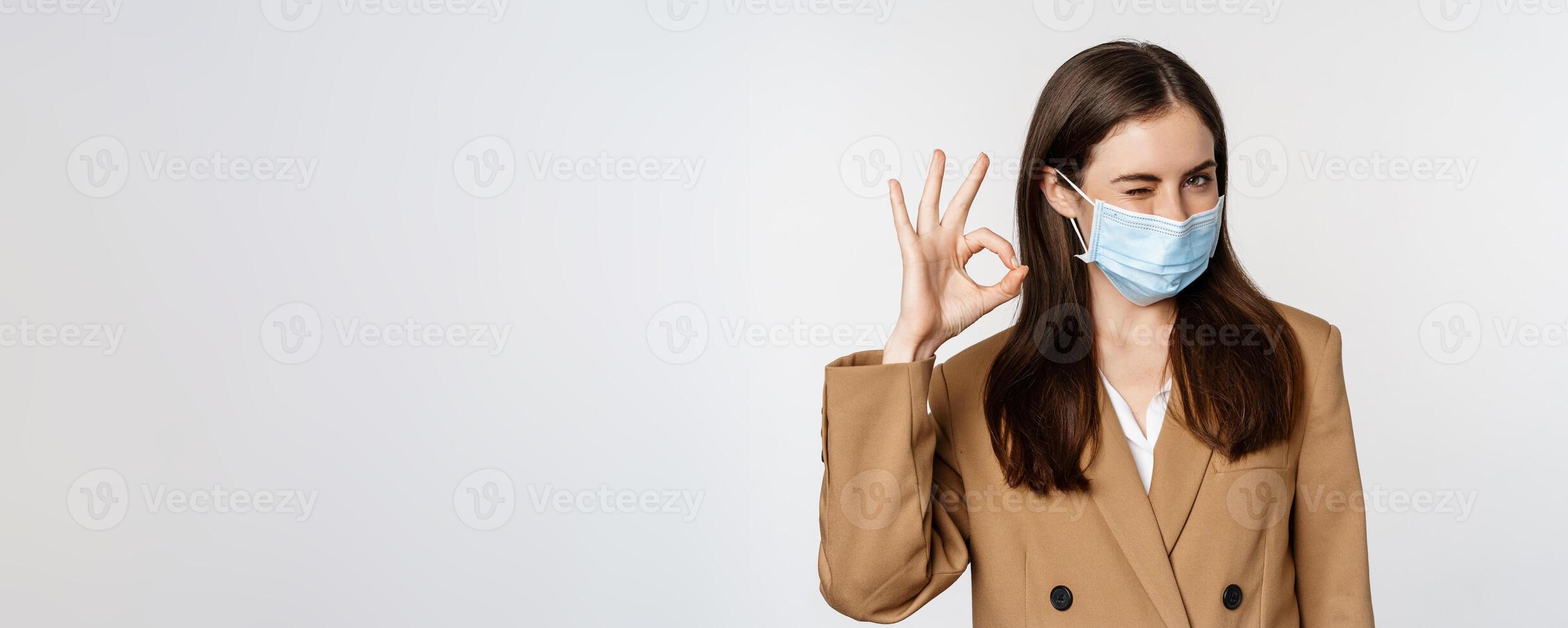 Workaplce and pandemic concept. Close up portrait of business woman showing okay signs, wearing medical mask, recommend, praise something, standing over white background photo