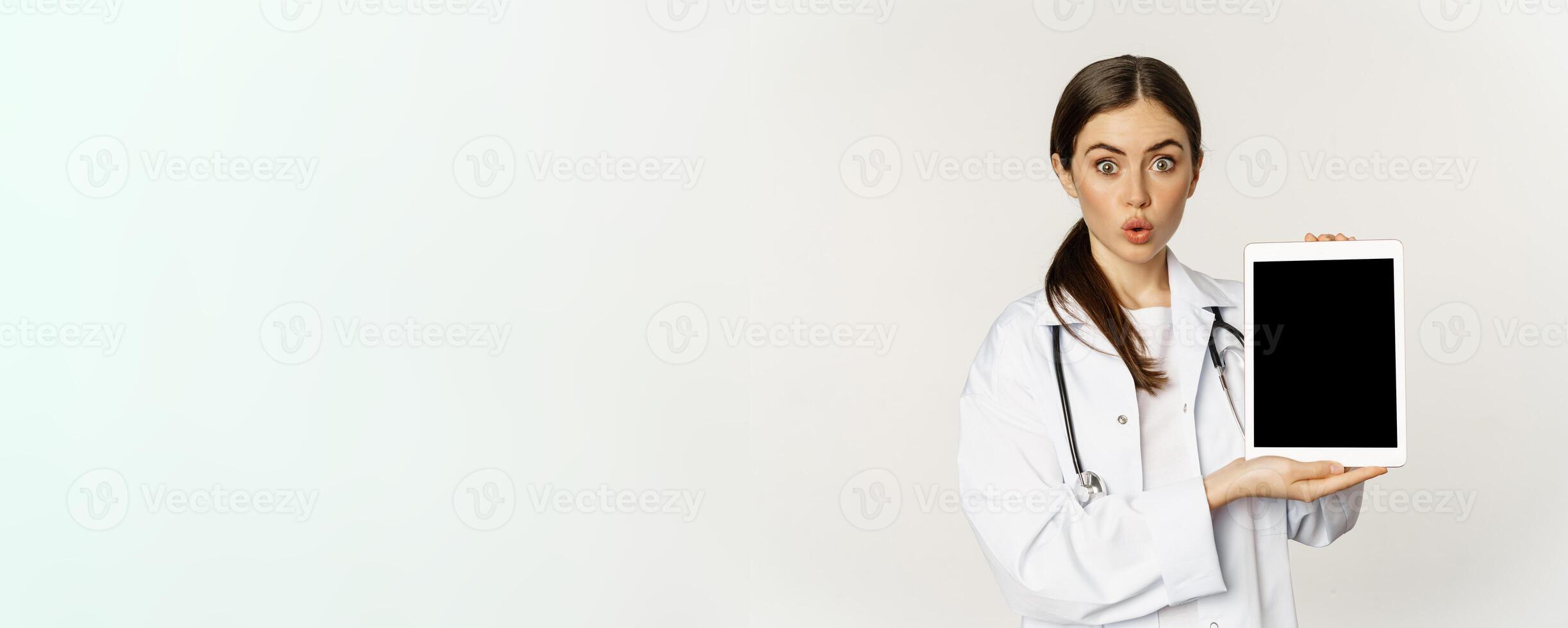 Image of woman doctor, female healthcare worker showing online medical website, digital tablet screen and smiling, standing in white coat over white background photo