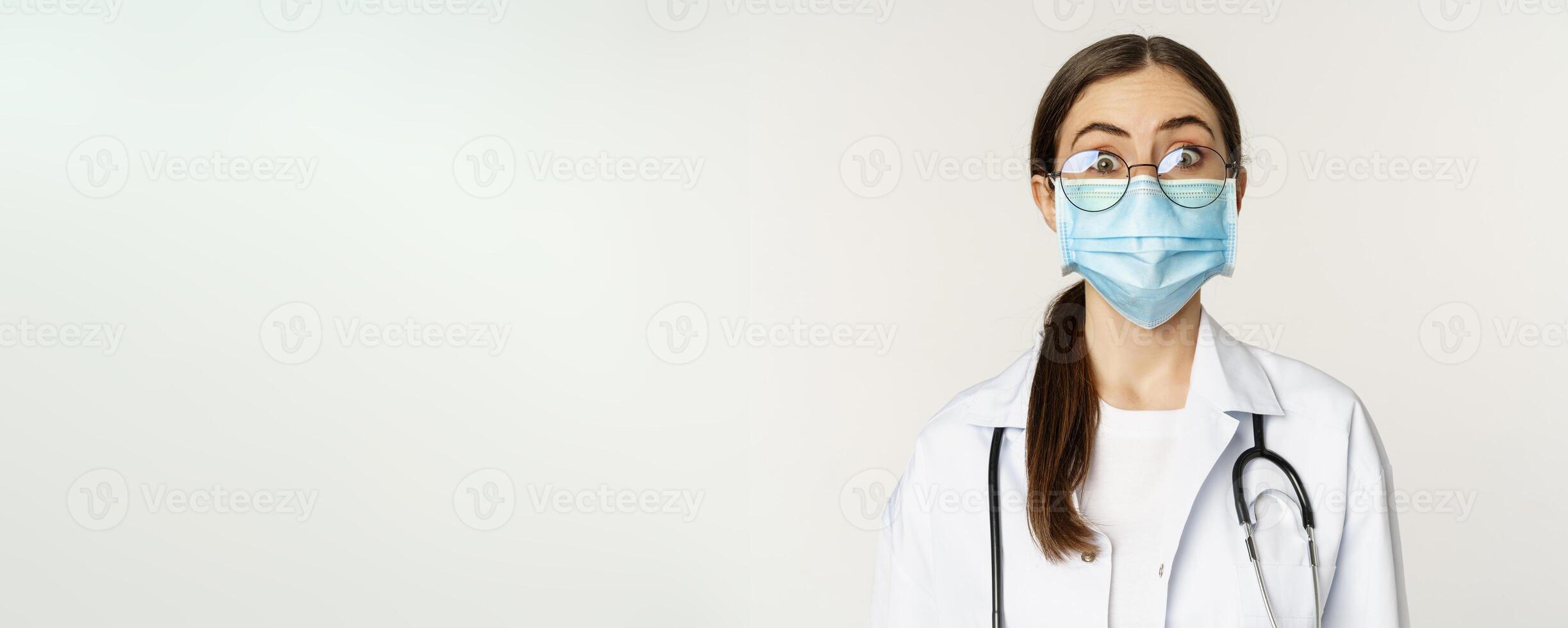 Medical worker, young doctor looking amazed and surprised, wearing face mask to prevent catching covid-19, standing over white background photo
