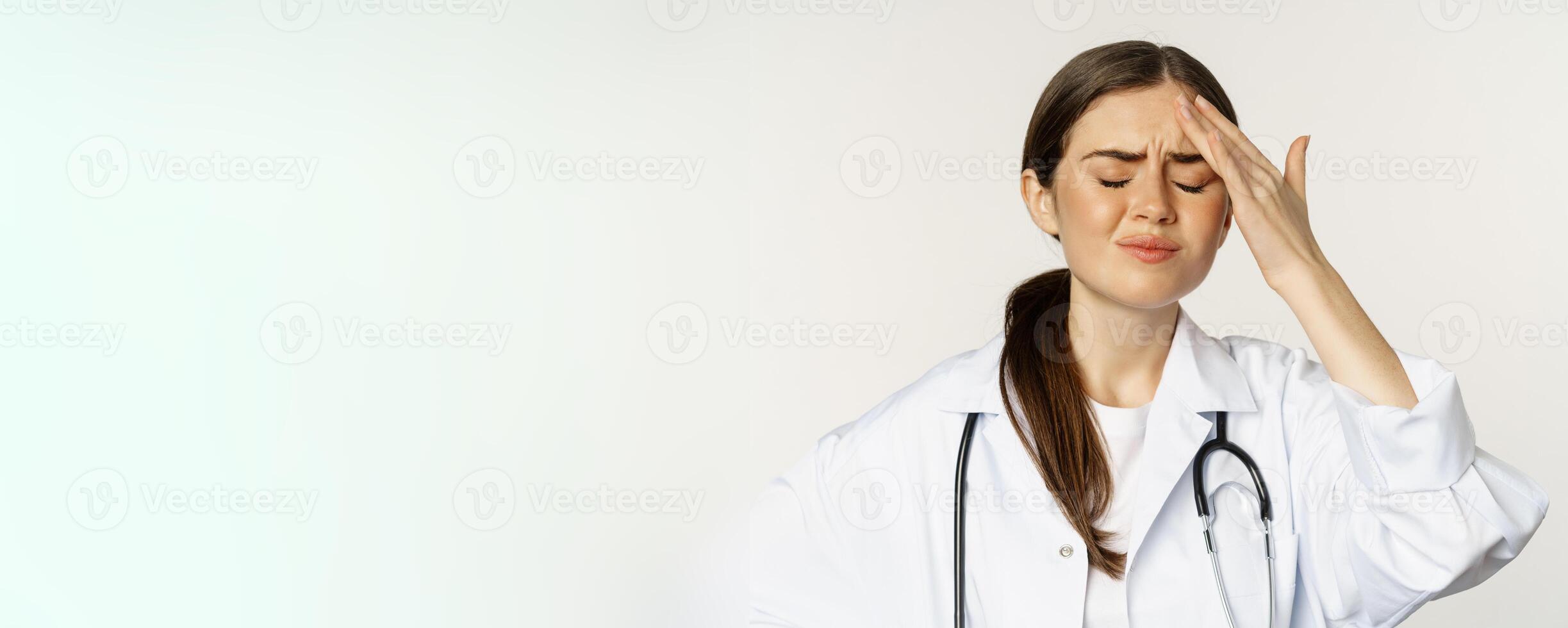Portrait of distressed, frustrated doctor, holding hands on forehead and grimacing from headache, painful migraine, standing over white background photo