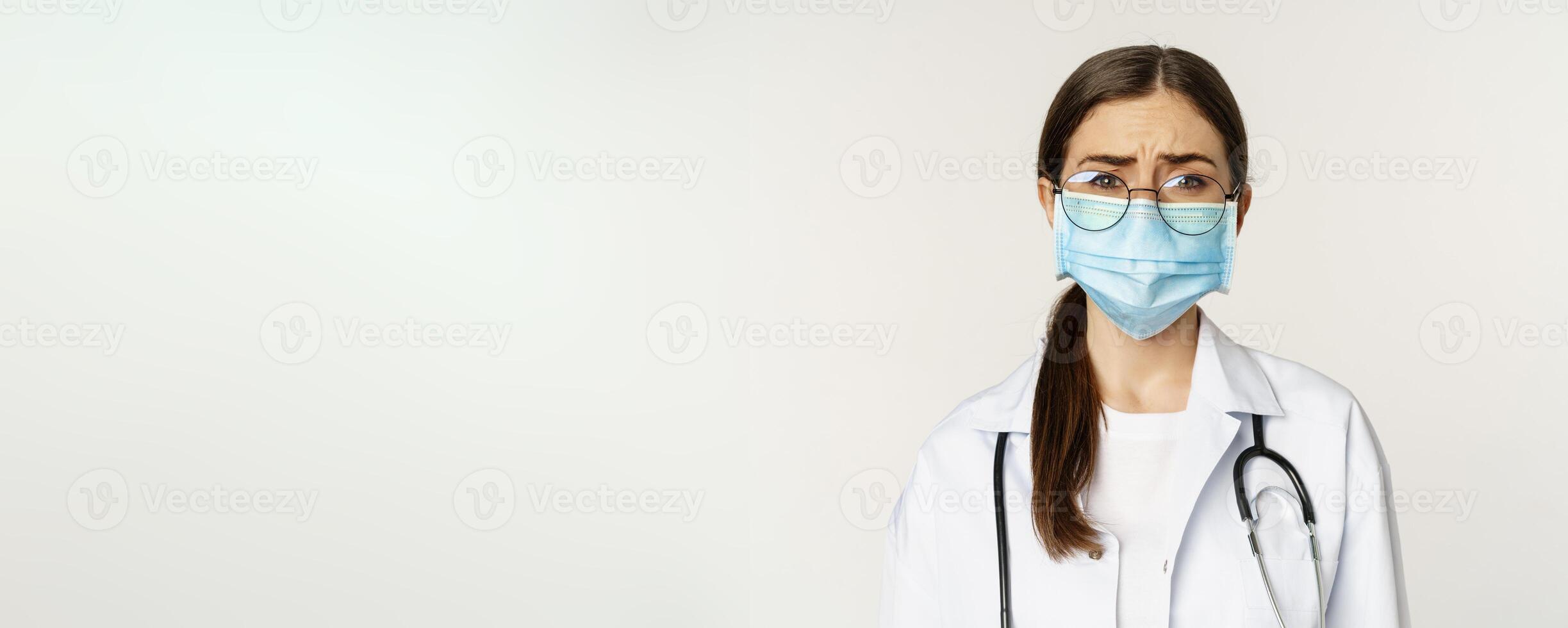 Sad and gloomy young woman doctor in medical face mask, feeling upset or concerned, frowning, standing over white background photo