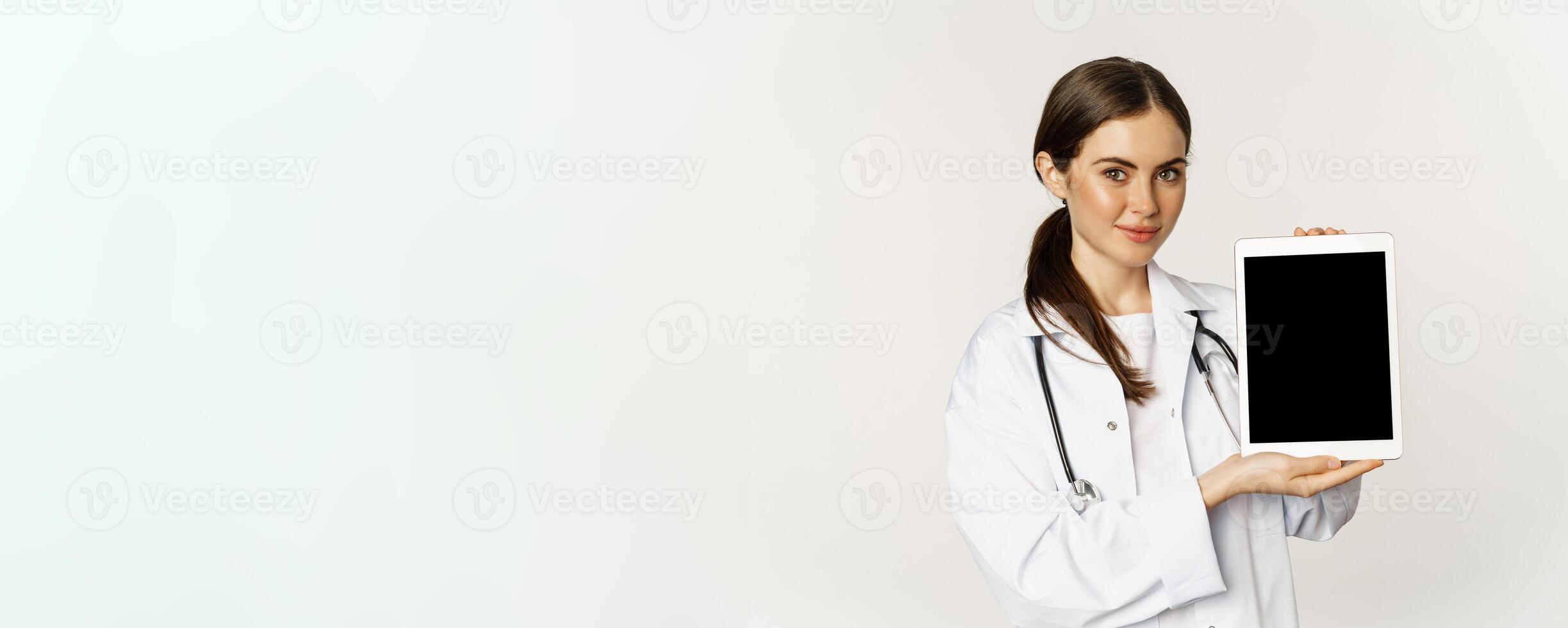 Image of woman doctor, female healthcare worker showing online medical website, digital tablet screen and smiling, standing in white coat over white background photo