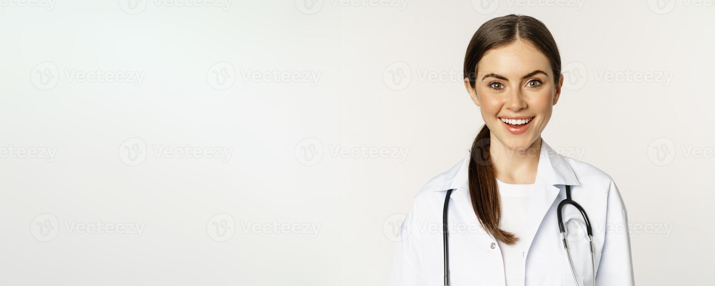 Smiling woman doctor, physician at appointment, looking happy and confident, wearing white coat and stethoscope, standing against studio background photo