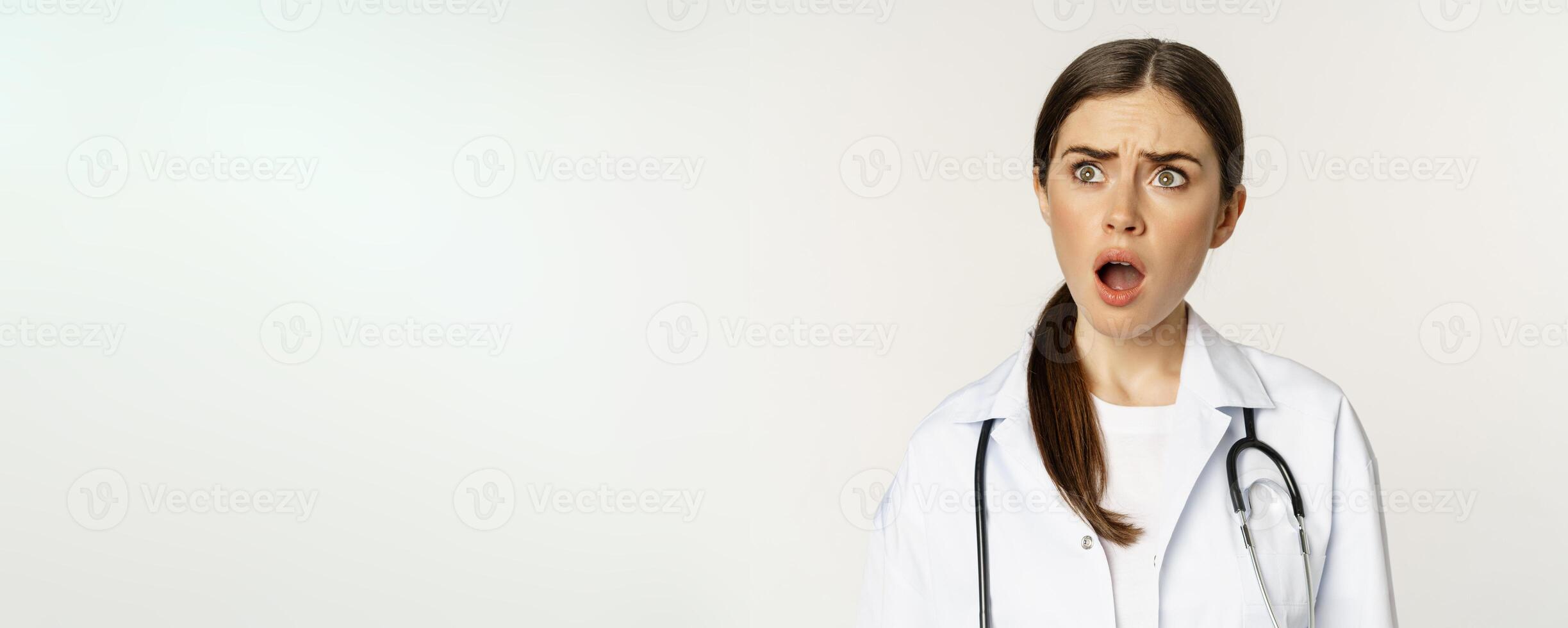 Frustrated and upset young woman, looking left with insulted shocked face, standing in doctor uniform over white background photo