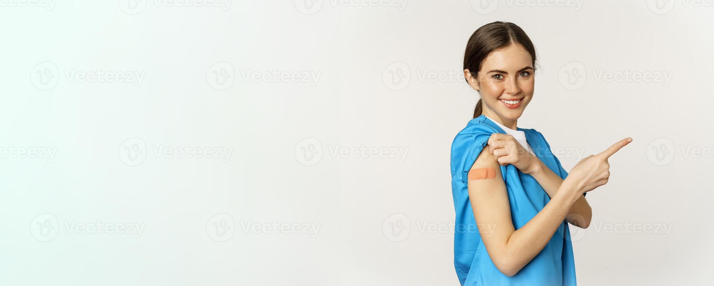Smiling nurse, female healthcare worker in scrubs, pointing finger right, showing patch on shoulder, vaccination campaign, covid-19 and pandemic concept, white background photo