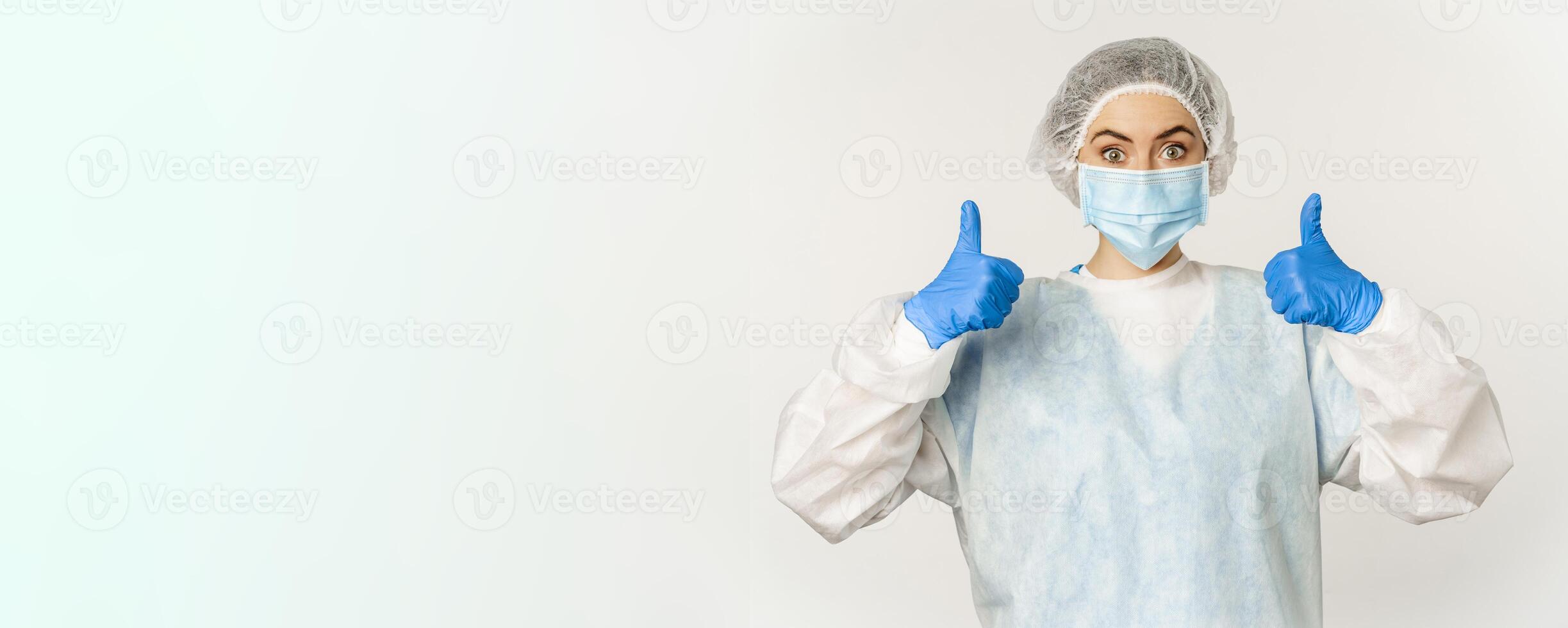 Portrait of doctor in personal protective equipment, showing thumbs up, wearing face mask, concept of covid-19 and pandemic, white background photo
