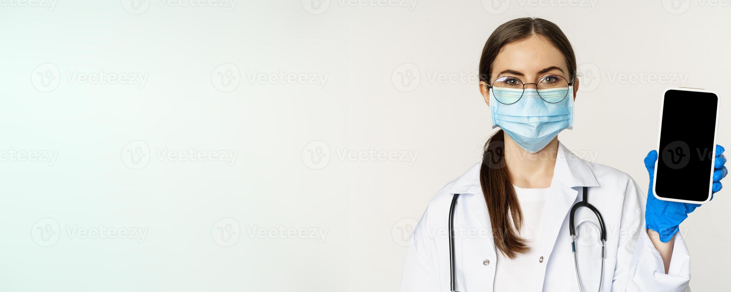 Online medical help concept. Woman doctor in glasses and face mask, showing mobile phone screen, app interface or website for patients, standing over white background photo