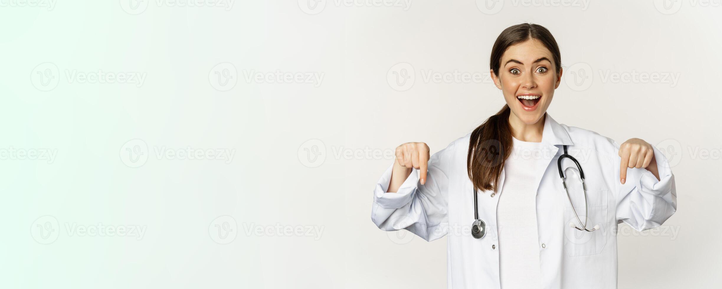 Portrait of happy female doctor, pointing fingers down and smiling, demonstrating promo offer, discount, standing over white background photo