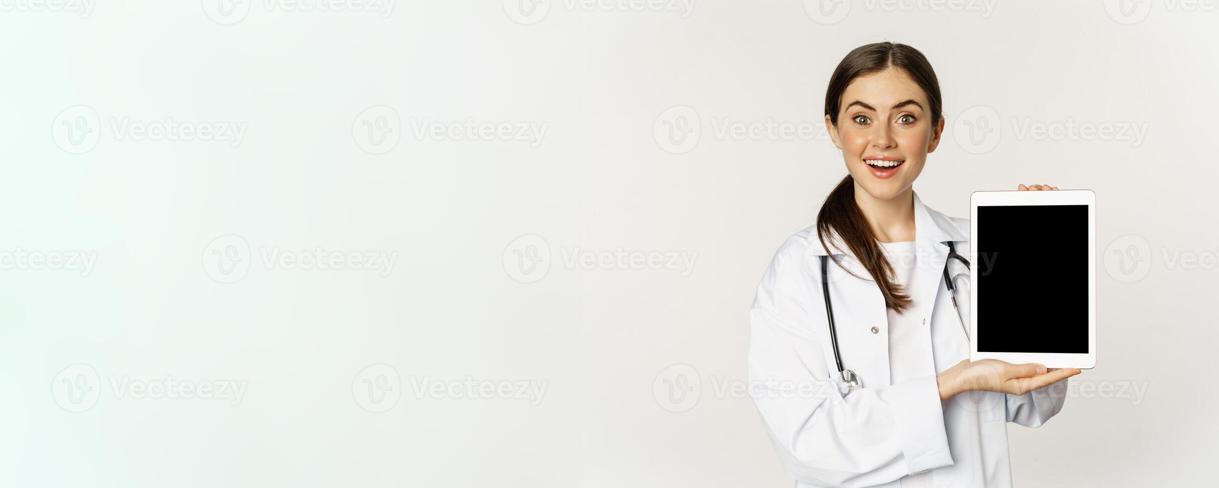 Image of woman doctor, female healthcare worker showing online medical website, digital tablet screen and smiling, standing in white coat over white background photo