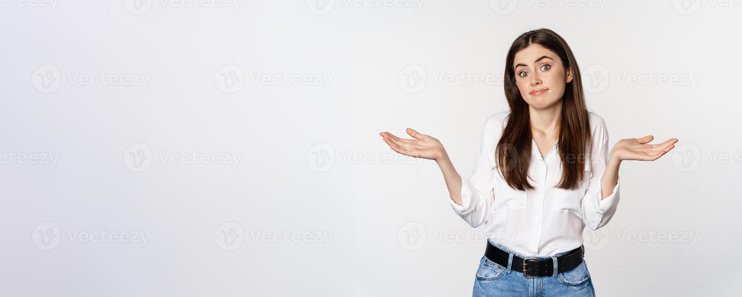 I dont know. Clueless young woman shrugging shoulders and looking unaware, standing over white background photo