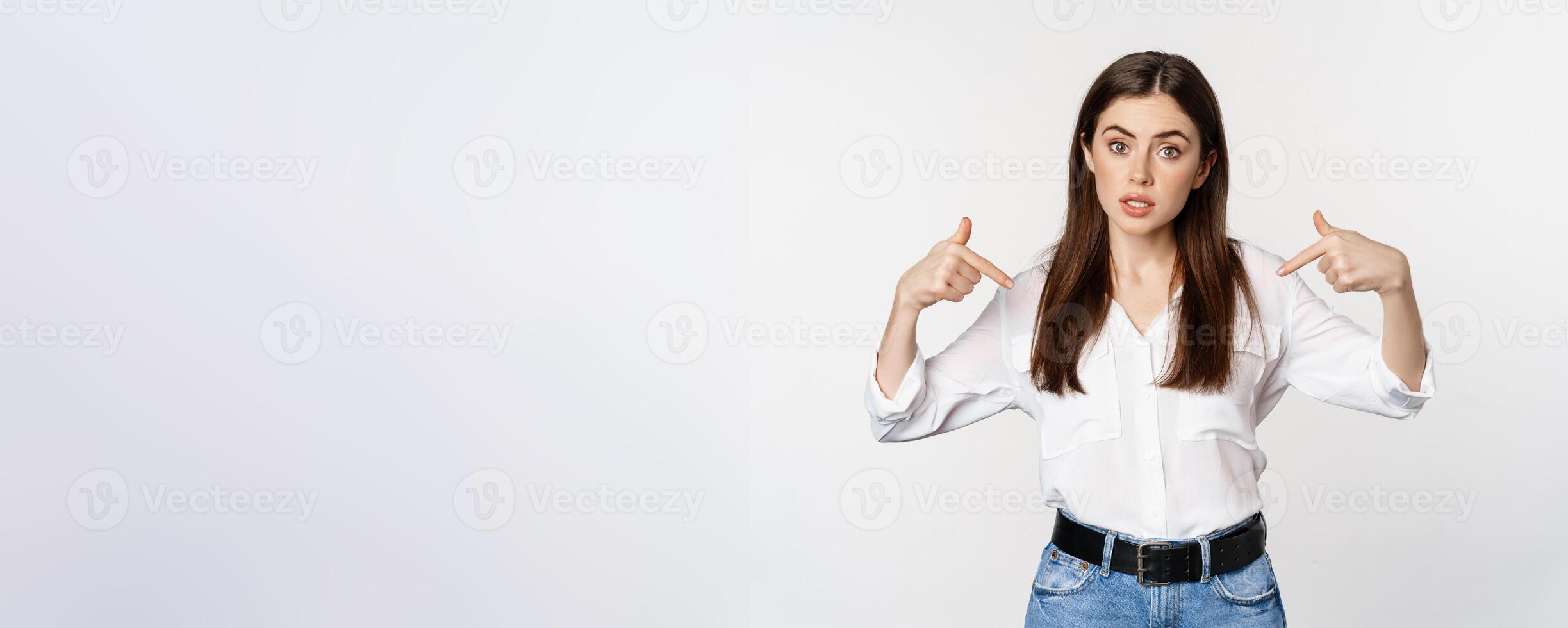 Portrait of young woman pointing at herself, self-promoting, standing over white background photo