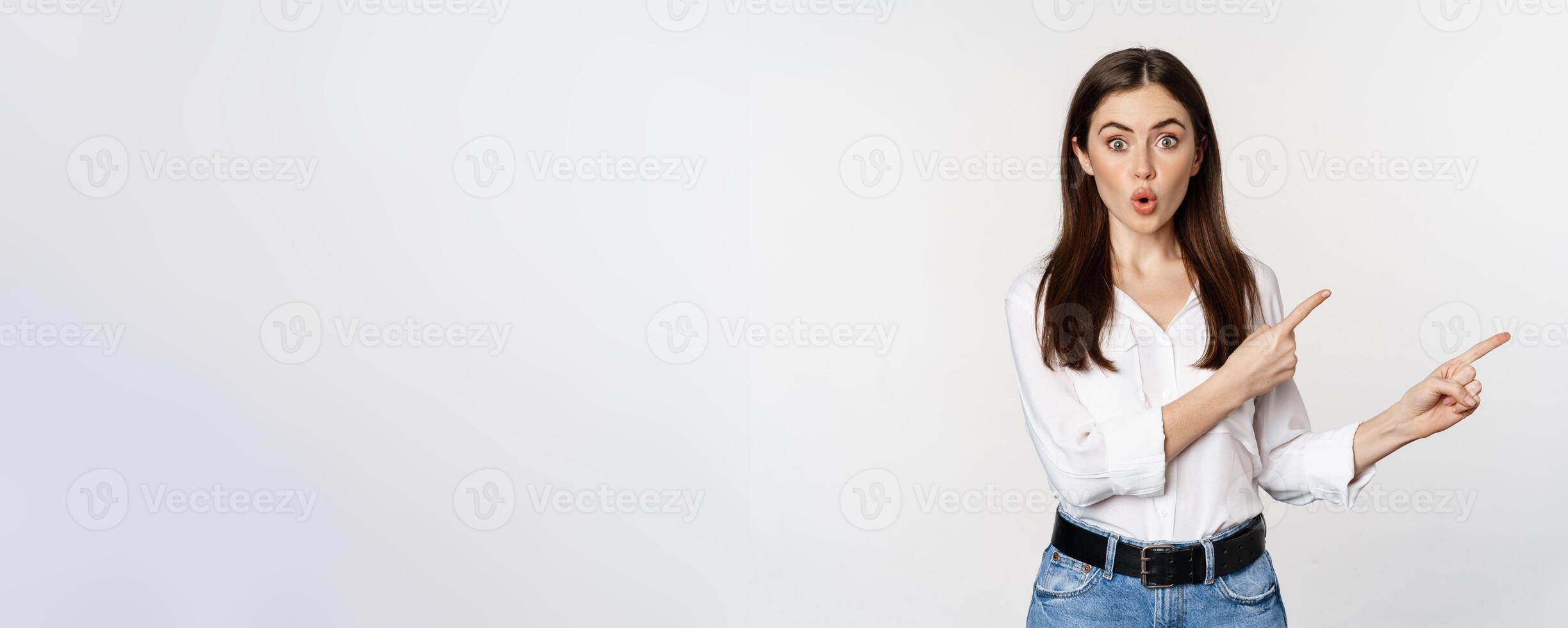 Amazed young woman pointing fingers down, showing announcement or logo banner, looking surprised and intrigued, standing over white background photo