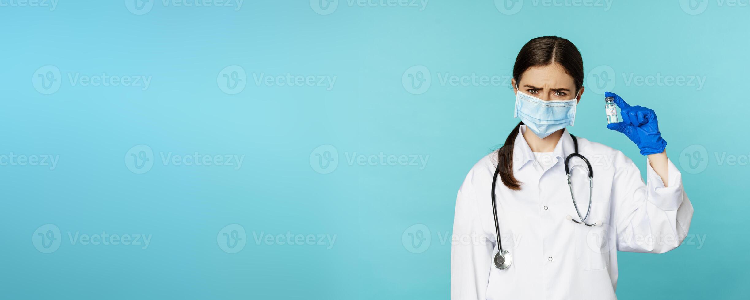 Upset and disappointed woman doctor in face medical mask, showing vaccine with sad dislike face, standing in uniform over blue background photo