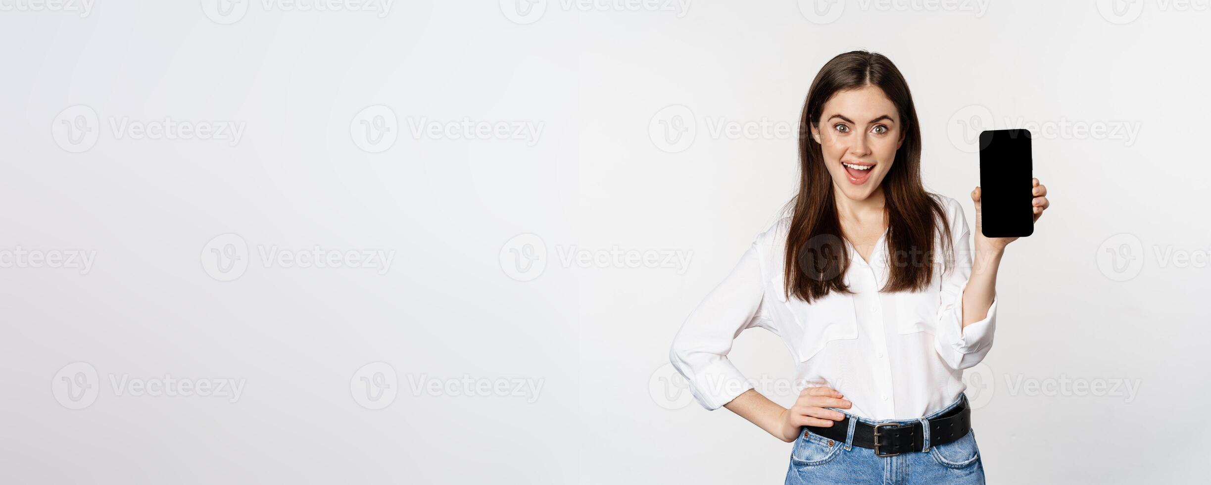 Happy smiling woman showing smartphone app interface, mobile phone screen, demonstrating promo offer, standing over white background photo