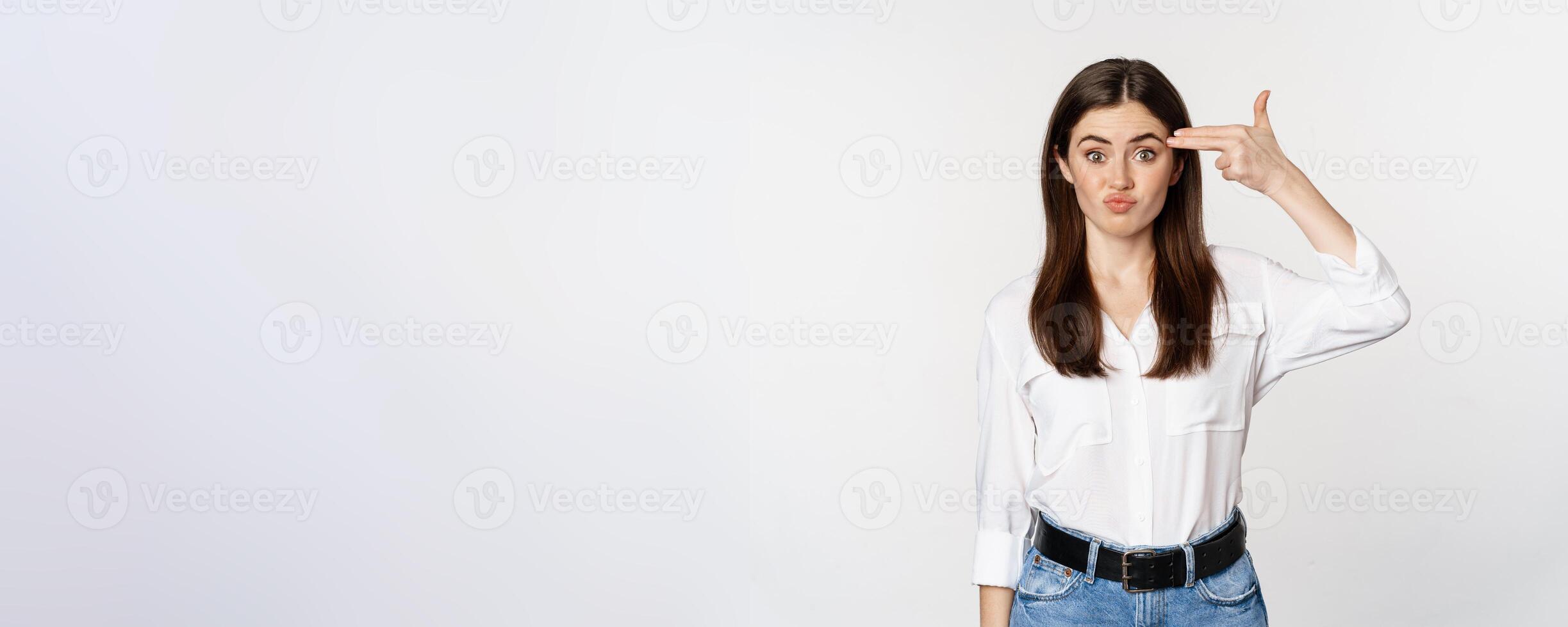 Image of annoyed young woman showing finger gun gesture over head, bothered by smth, tired, standing over white background photo