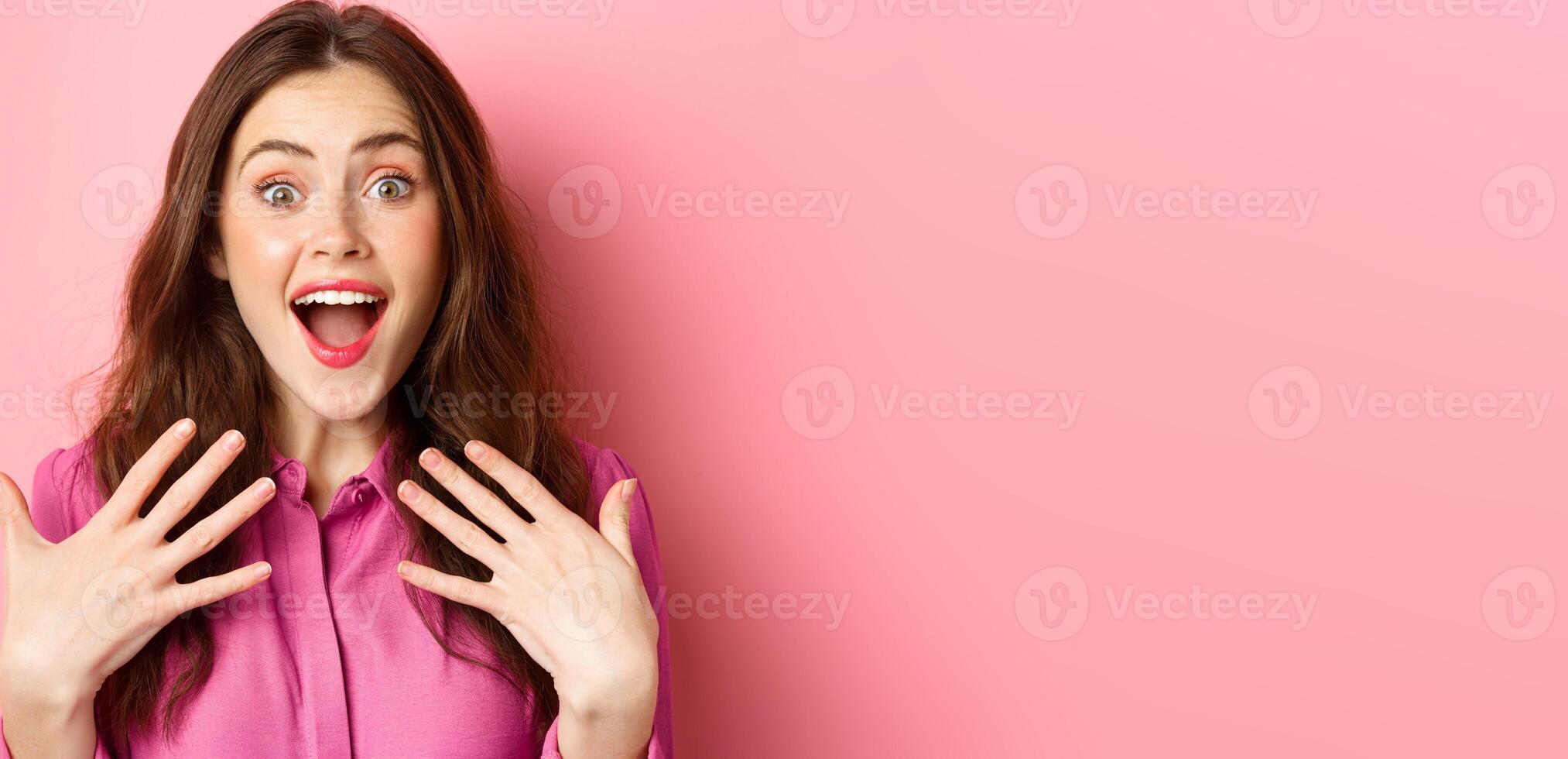 Close up of grateful and excited young woman gasping, holding hands on chest and stare with disbelief, receive awesome gift, standing against pink background photo