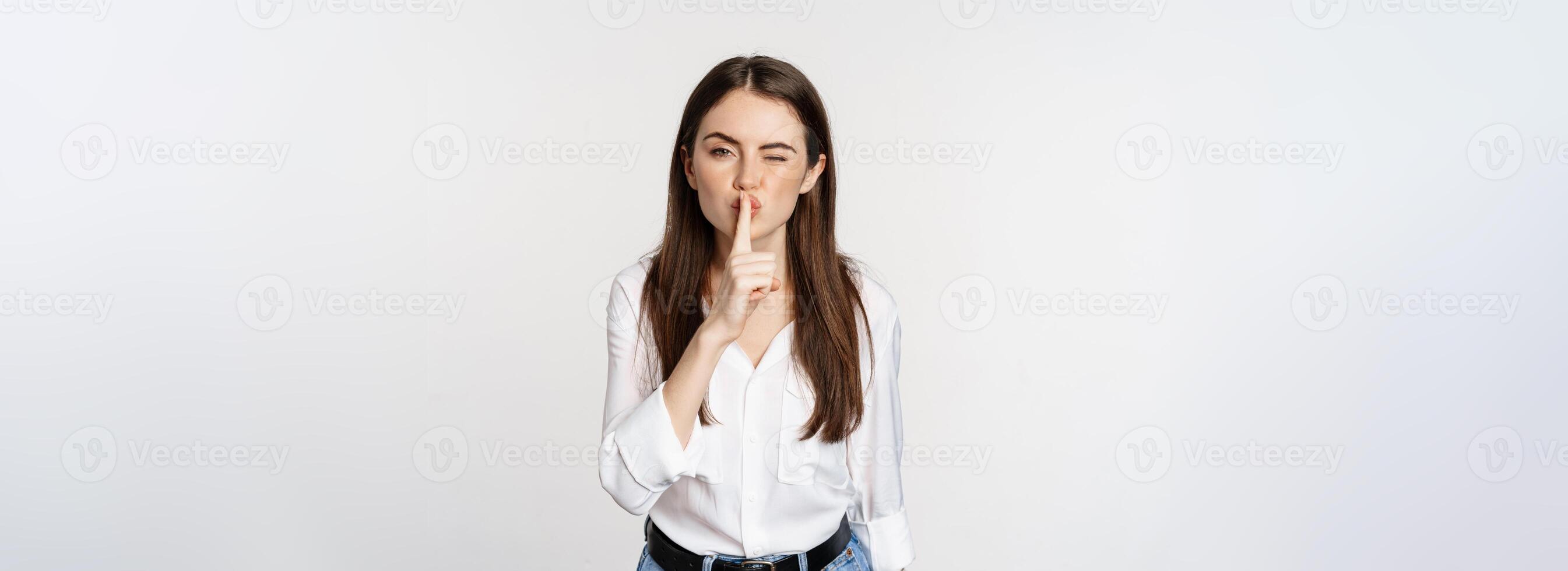 Smiling beautiful woman shushing, making hush shh sign, press finger to lips and winking, hiding secret, standing in blouse and jeans over white background photo