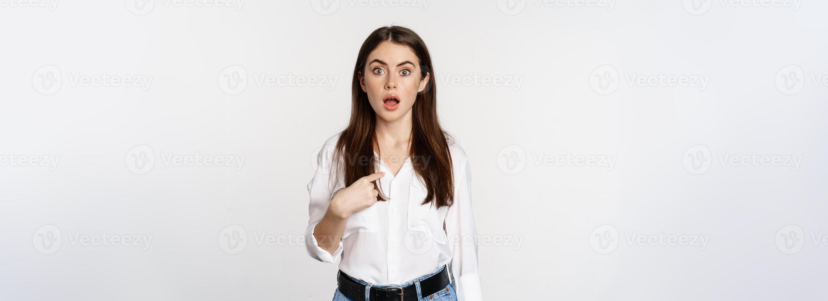 Portrait of young woman pointing at herself with disbelief, surprised face expression, being named or chosen, standing over white background photo