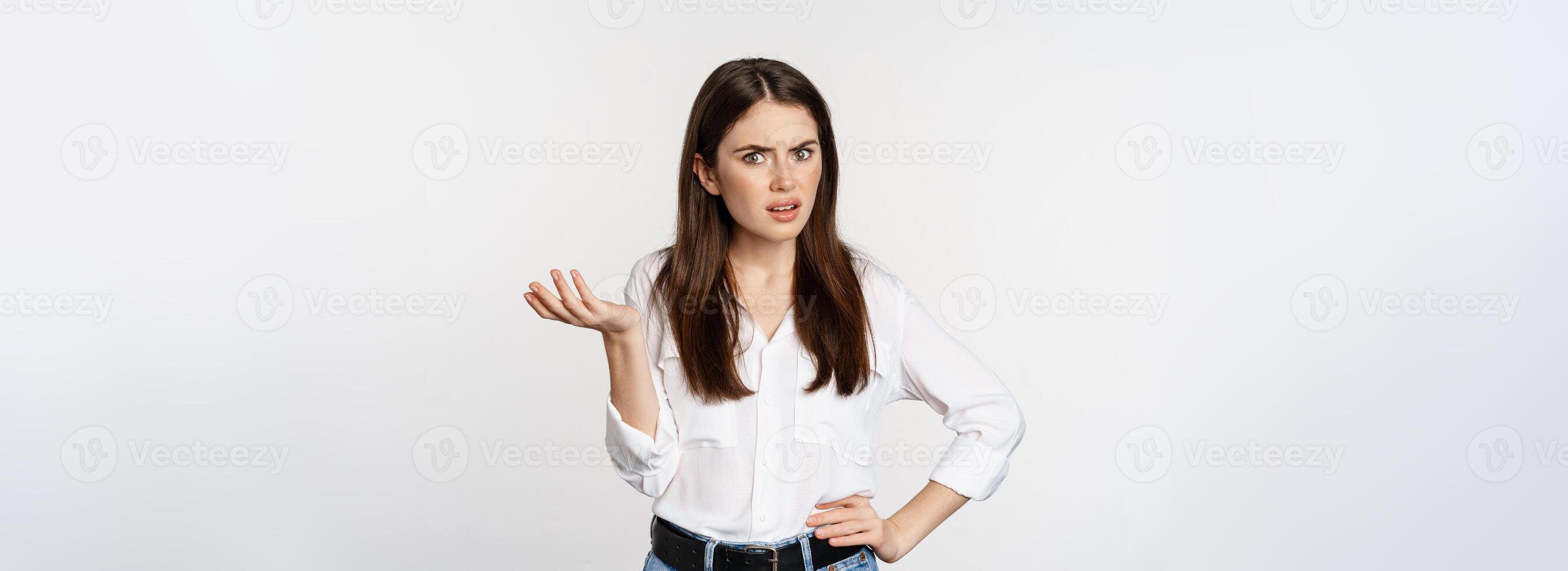 Confused business woman looking clueless and annoyed, cant understand smth strange, shaking hand and shrugging, standing over white background photo