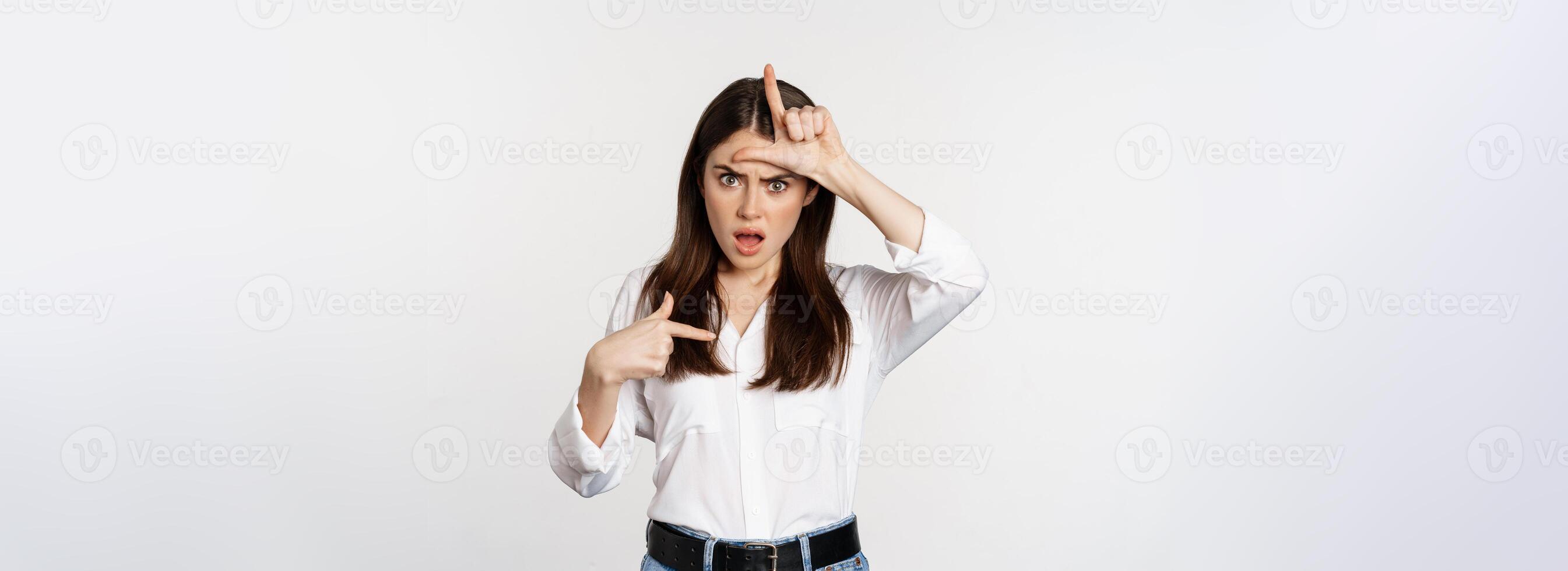 Insulted woman showing loser L word sign on forehead, pointing at herself frustrated, standing over white background photo