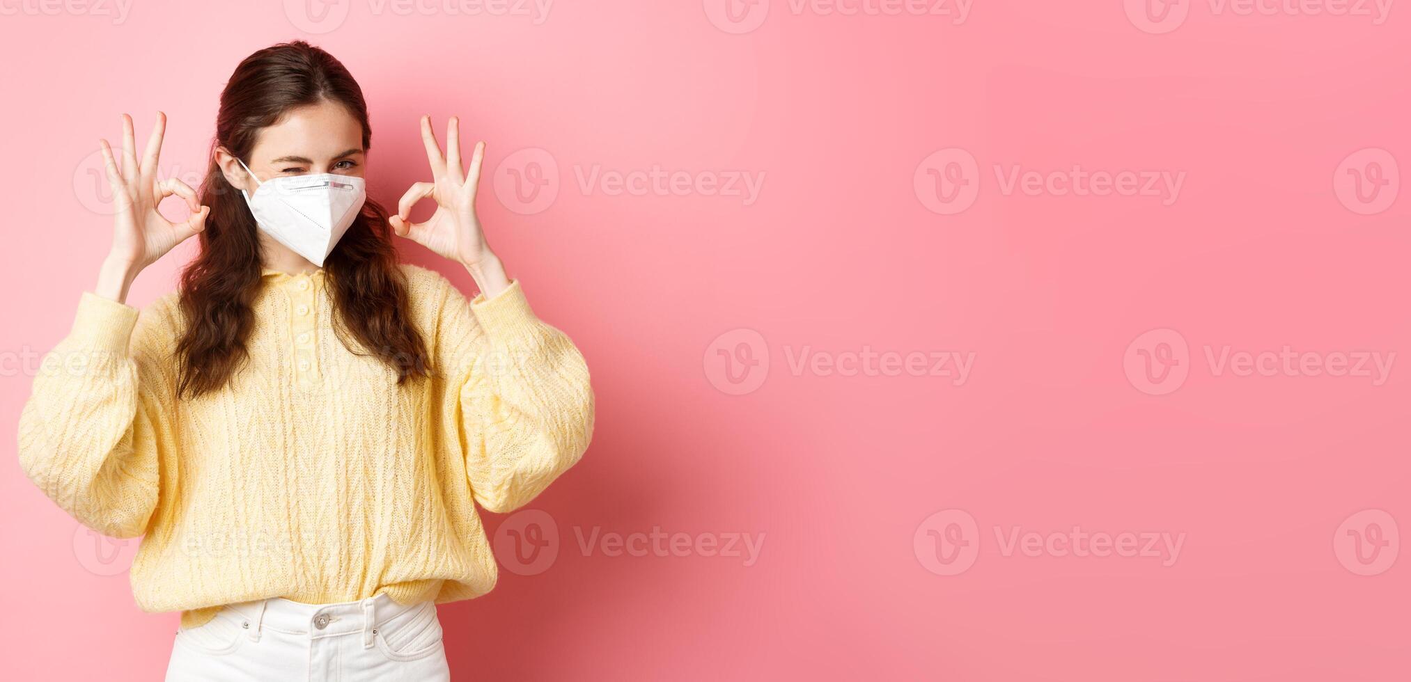 Covid-19, lockdown and pandemic concept. Young smiling girl assure you, winking and showing okay sign, alright or approve gesture, wearing medical respirator, standing over pink background photo