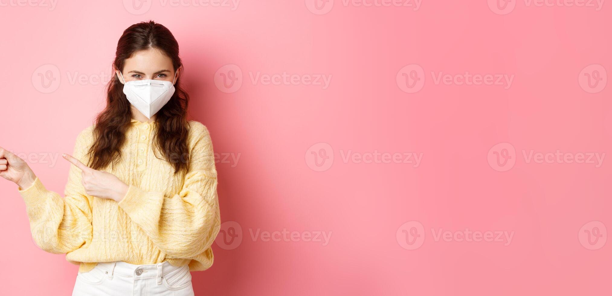 Covid, corona and social distancing concept. Coy smiling girl squinting thoughtful, pointing fingers aside at logo banner, wearing medical respirator during quarantine, pink background photo