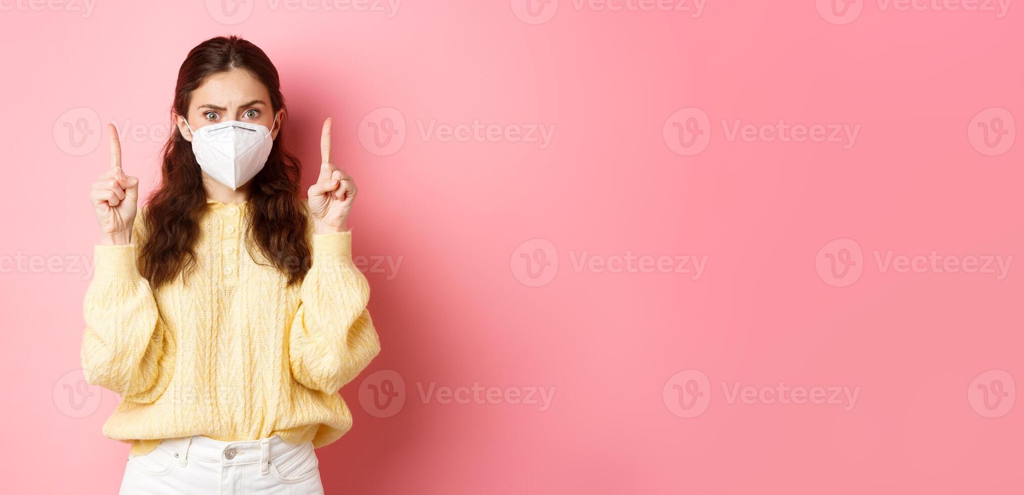 Covid, corona and social distancing concept. Confused and shocked young woman in medical respirator pointing fingers up, staring startled at camera, pink background photo