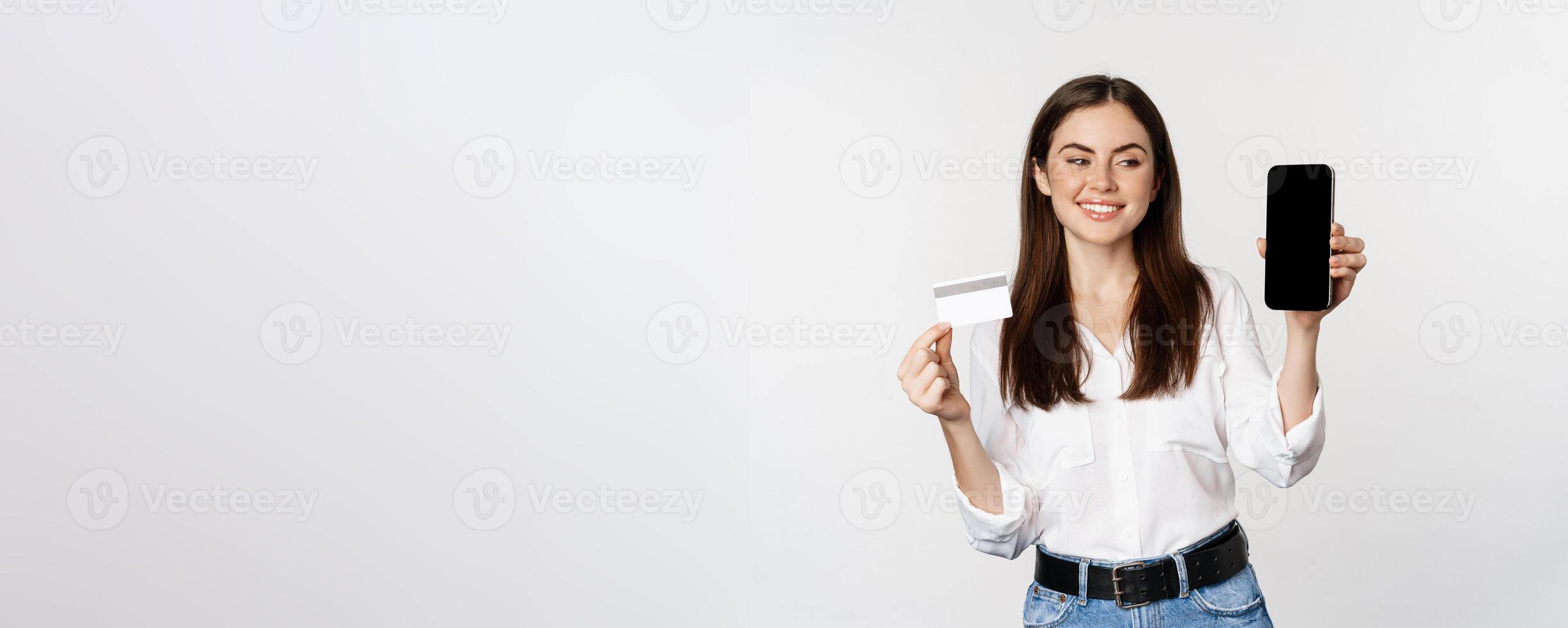 Portrait of female model showing credit card with smartphone screen, recommending application, standing over white background photo