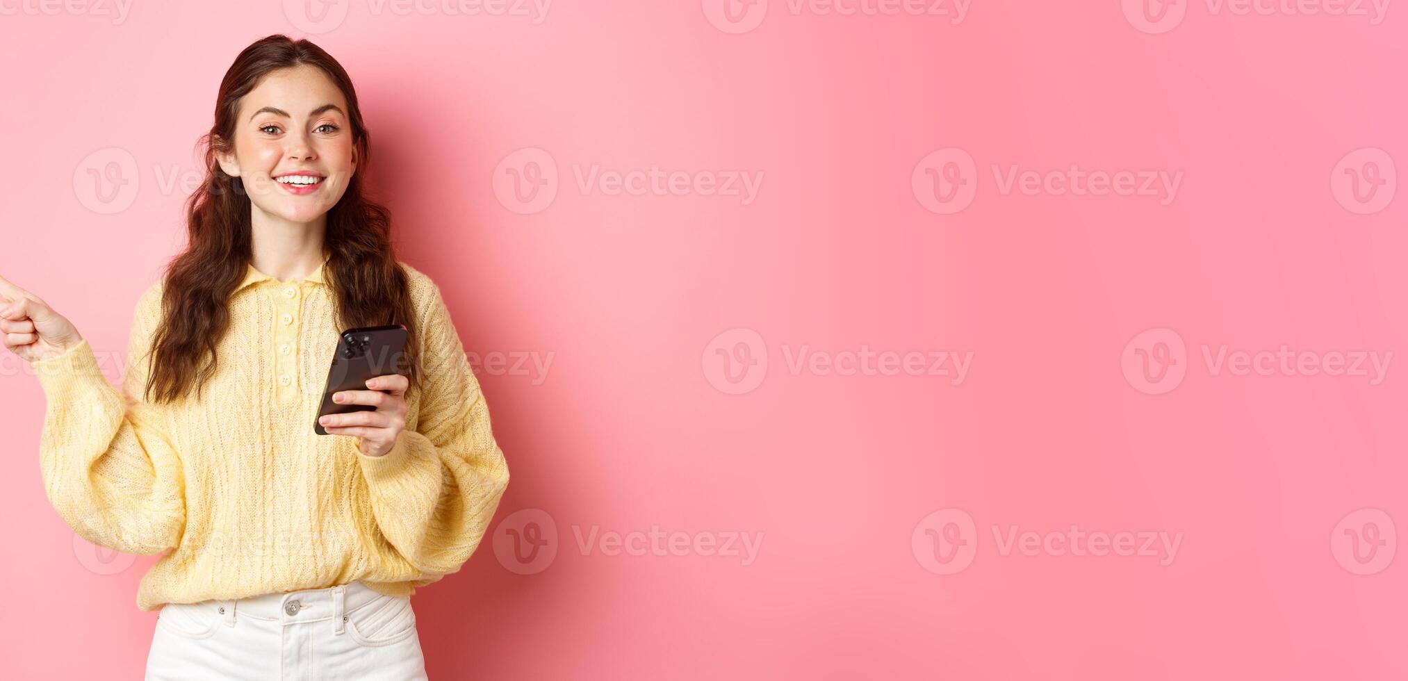 Beautiful modern girl holding smartphone and pointing finger aside, showing promotional text on copyspace while using mobile phone, standing over pink background photo