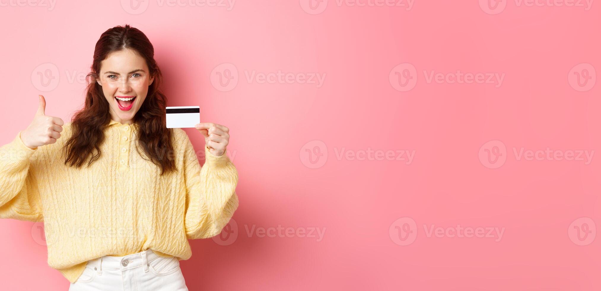Young modern woman saying yes and showing thumbs up, holding plastic credit card, standing over pink background photo