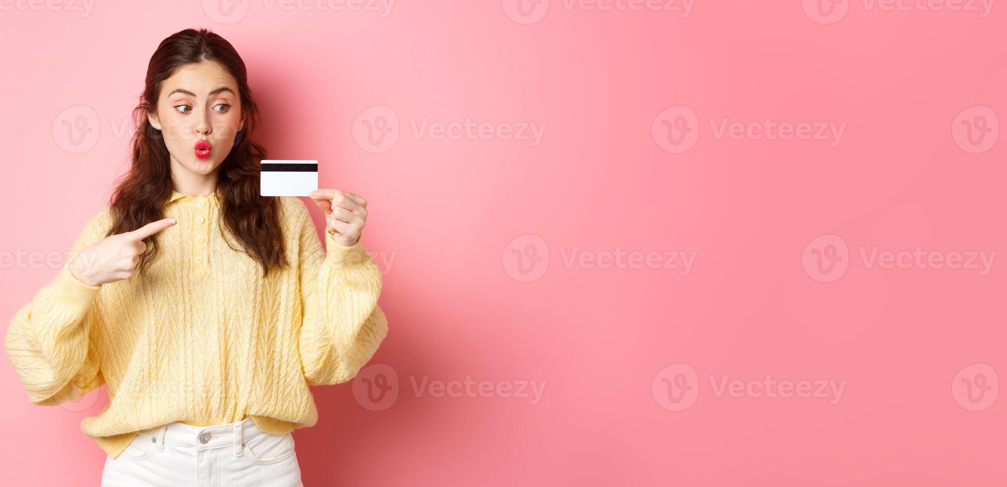 Image of glamour girl going shopping, looking excited and pointing at plastic credit card, standing against pink background photo