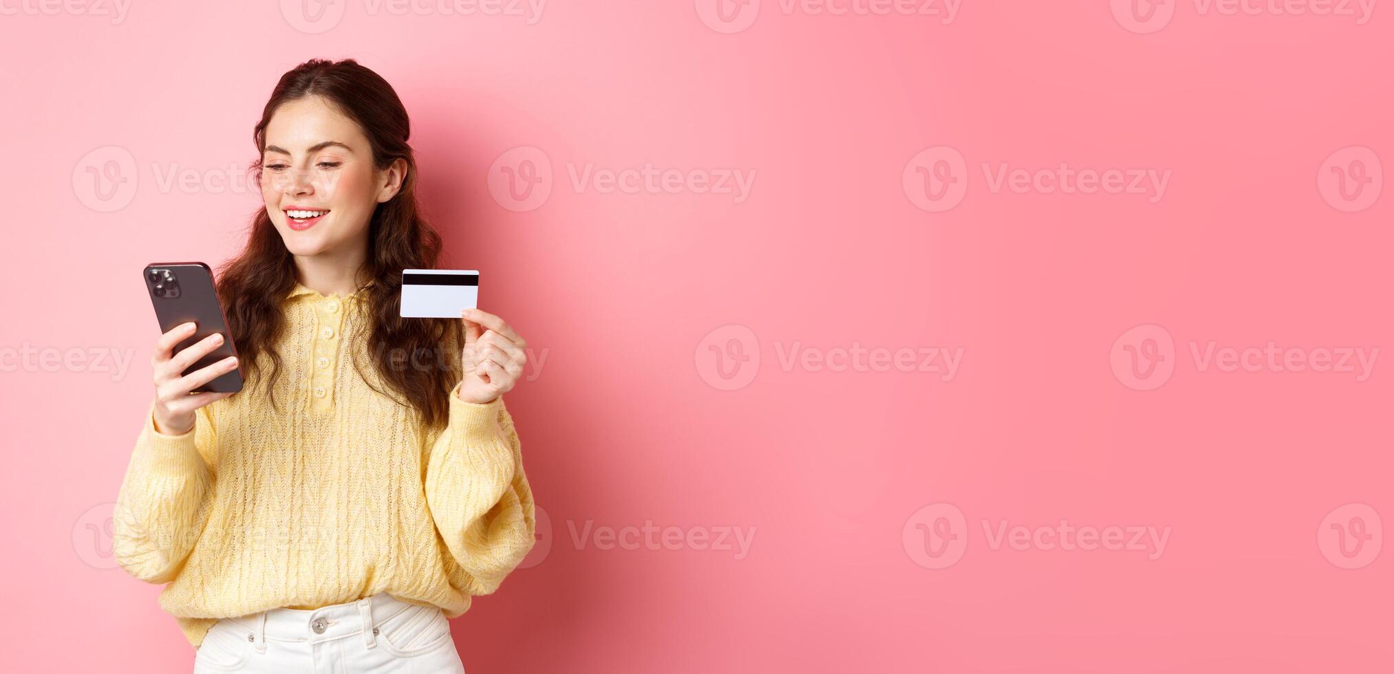 Technology and online shopping. Young pretty lady paying online with credit card, looking at smartphone and smiling, standing over pink background photo