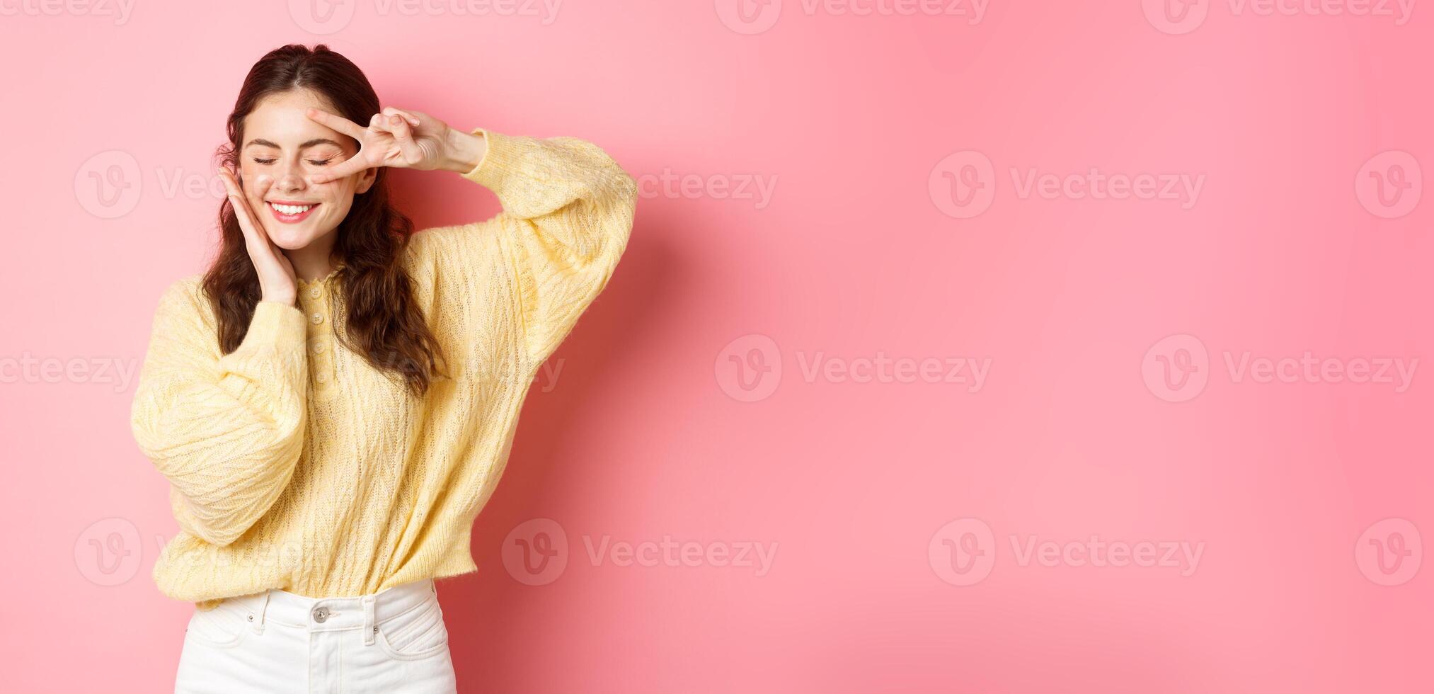 Image of beautiful brunette female model, smiling and touching her face, showing v-sign near eye, laughing happy, standing over pink background photo