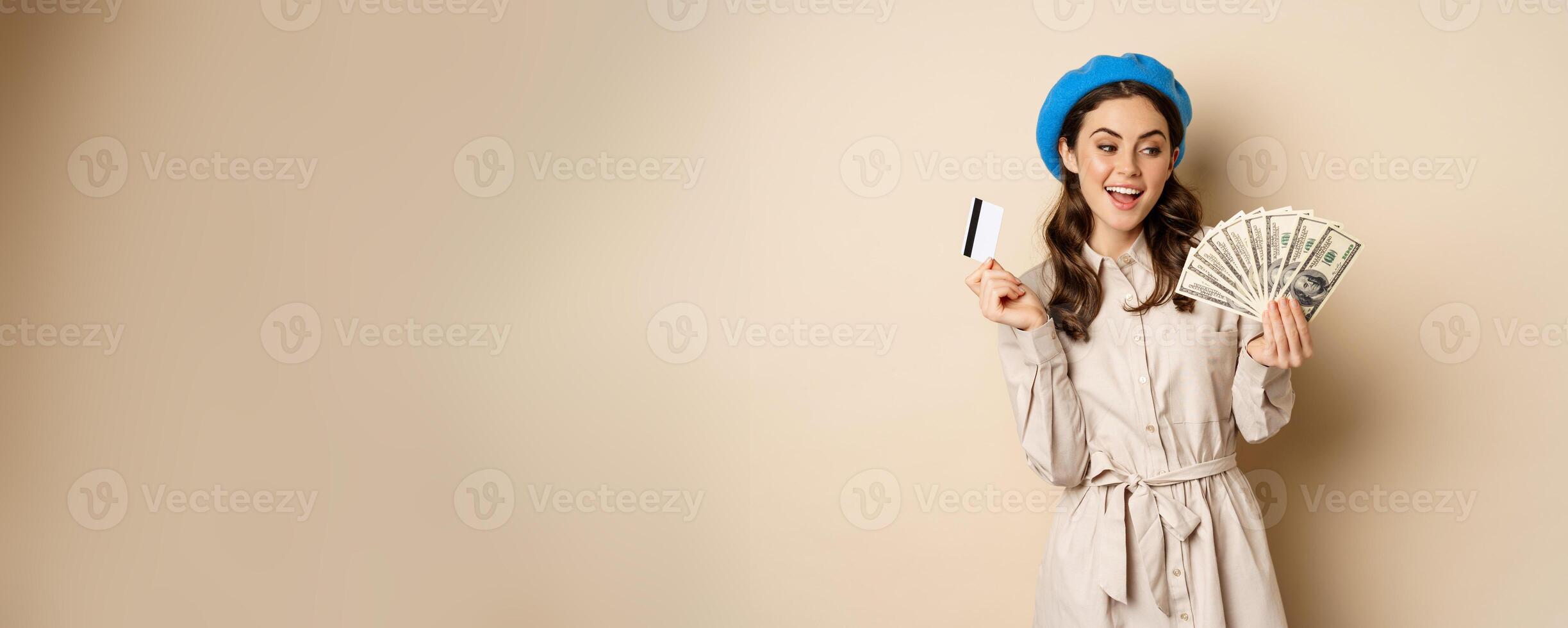 Microcredit and money concept. Young stylish woman showing credit card and dollars cash, smiling happy and satisfied, standing over beige background photo
